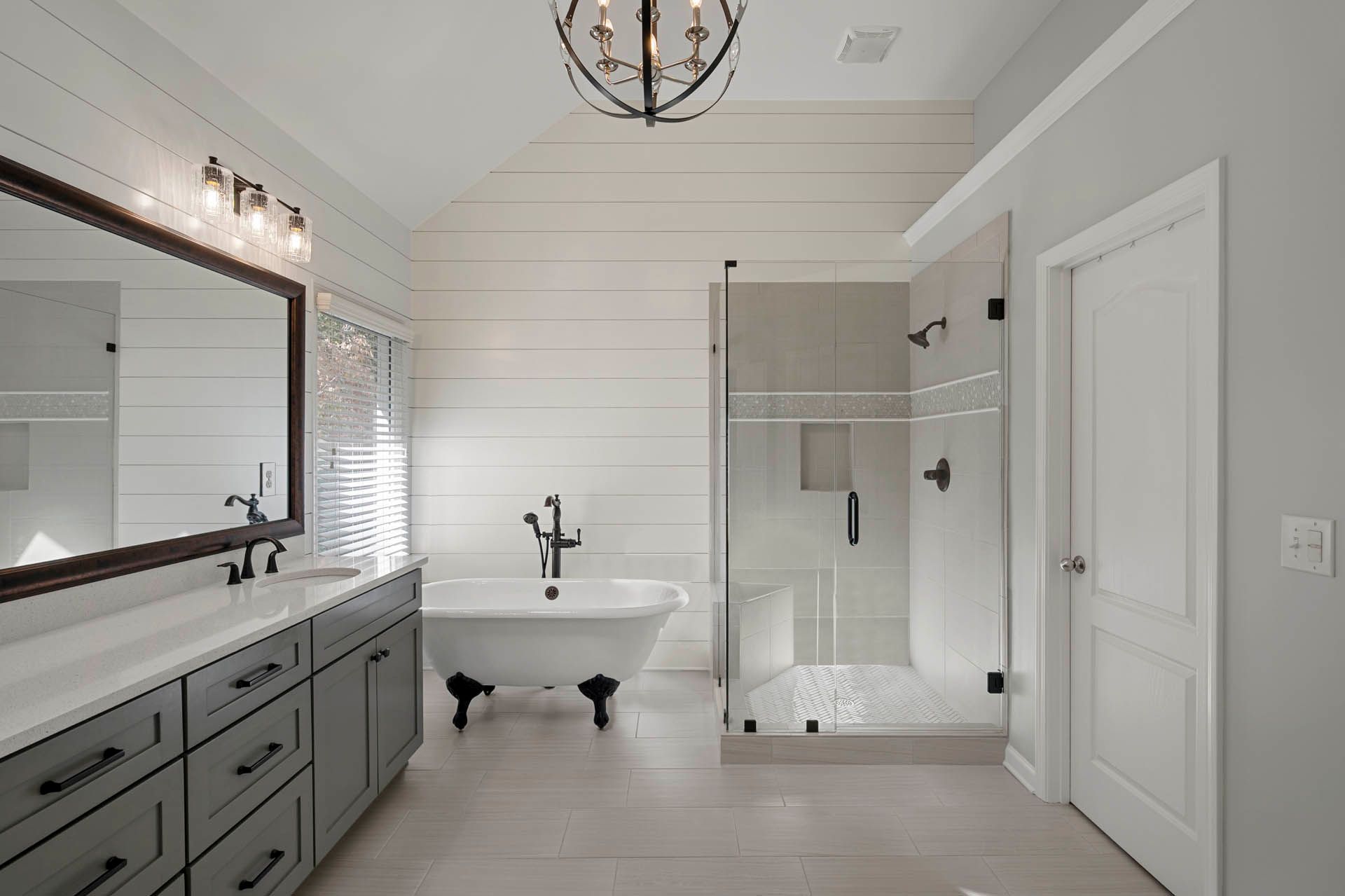 Bathroom with white walls, gray cabinets, a clawfoot tub, and a glass shower. A large mirror and chandelier are also present.