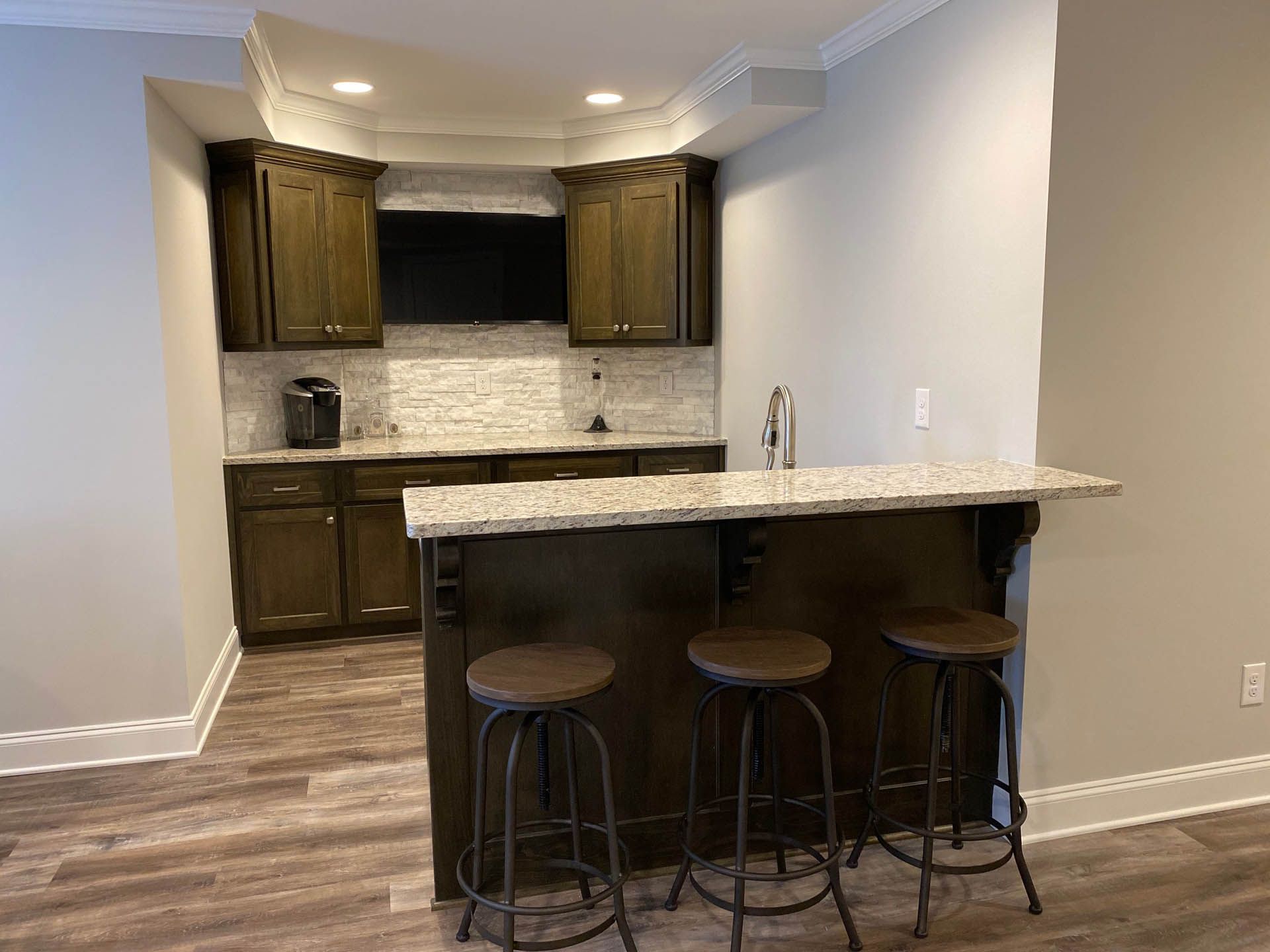A basement bar with dark brown cabinets, a granite countertop, and three stools. A small TV is mounted above the cabinets.