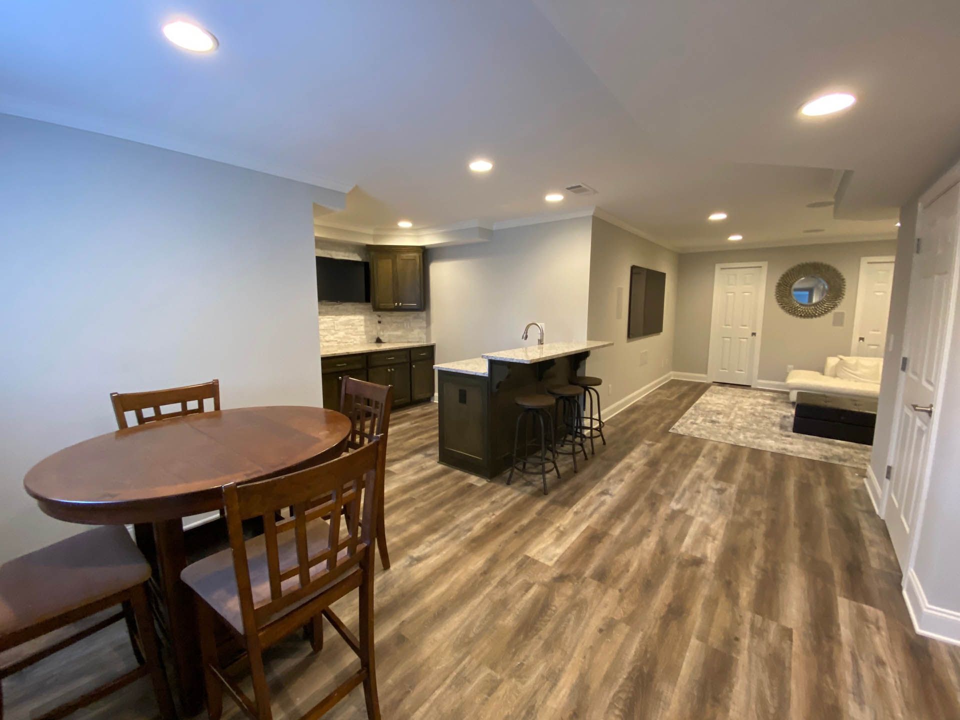 Basement living space with a dining table, kitchen, and seating area. Gray walls, wood-look flooring, dark cabinets.