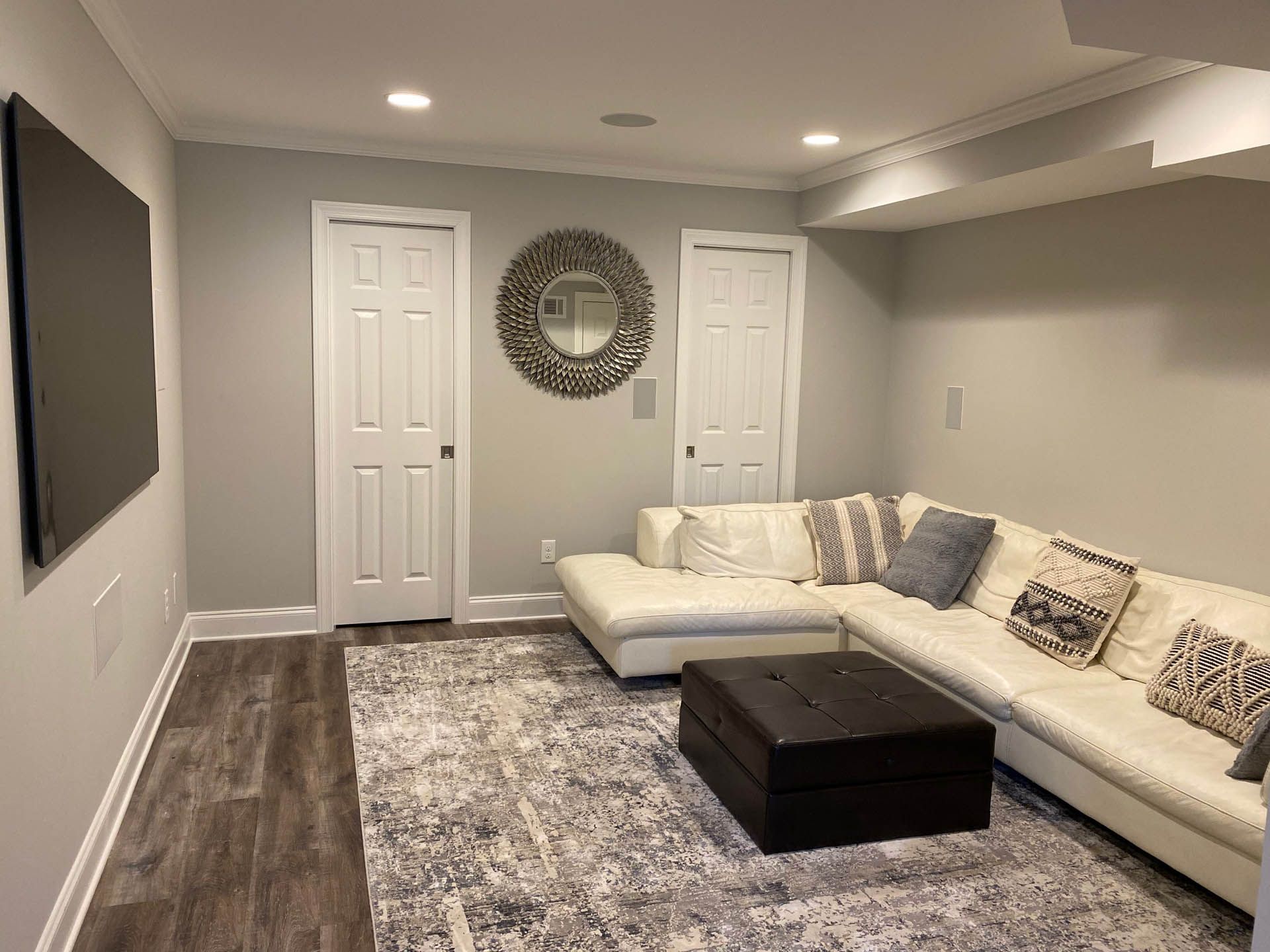 A modern living room with a white sectional sofa, area rug, and a large screen on the wall. Two white doors and a round mirror are visible.