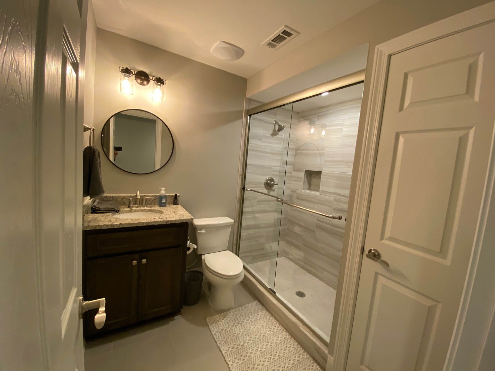 Bathroom with a dark wood vanity, toilet, and a shower with a glass door and white tiled walls. A round mirror hangs above the sink.