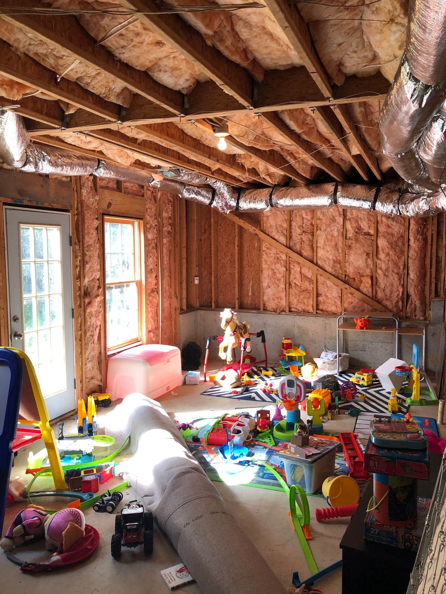 Unfinished basement playroom with exposed beams and insulation. Toys are scattered across the floor.