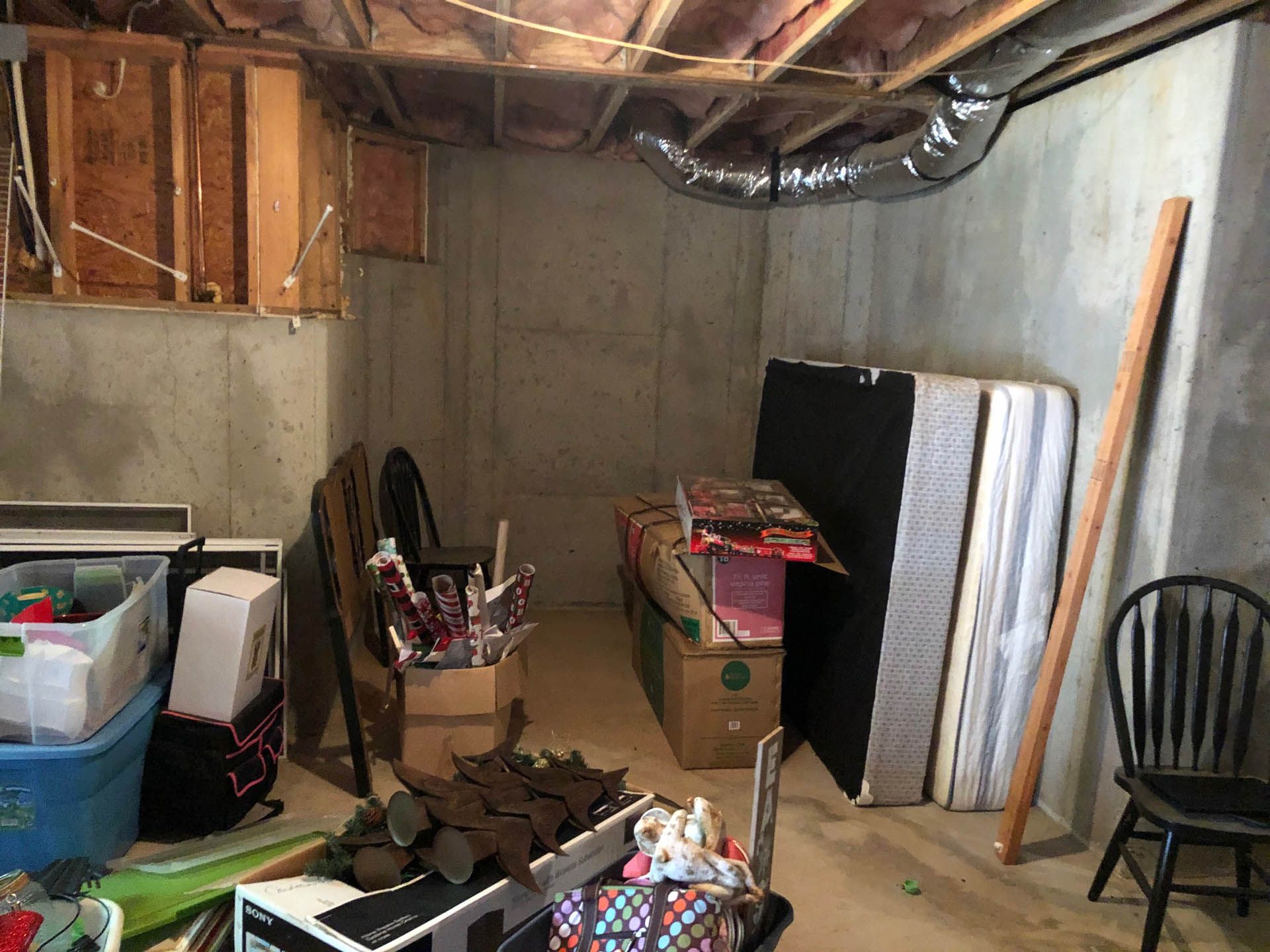 Basement with concrete walls, boxes of items, mattress leaning against a wall, and a black chair.