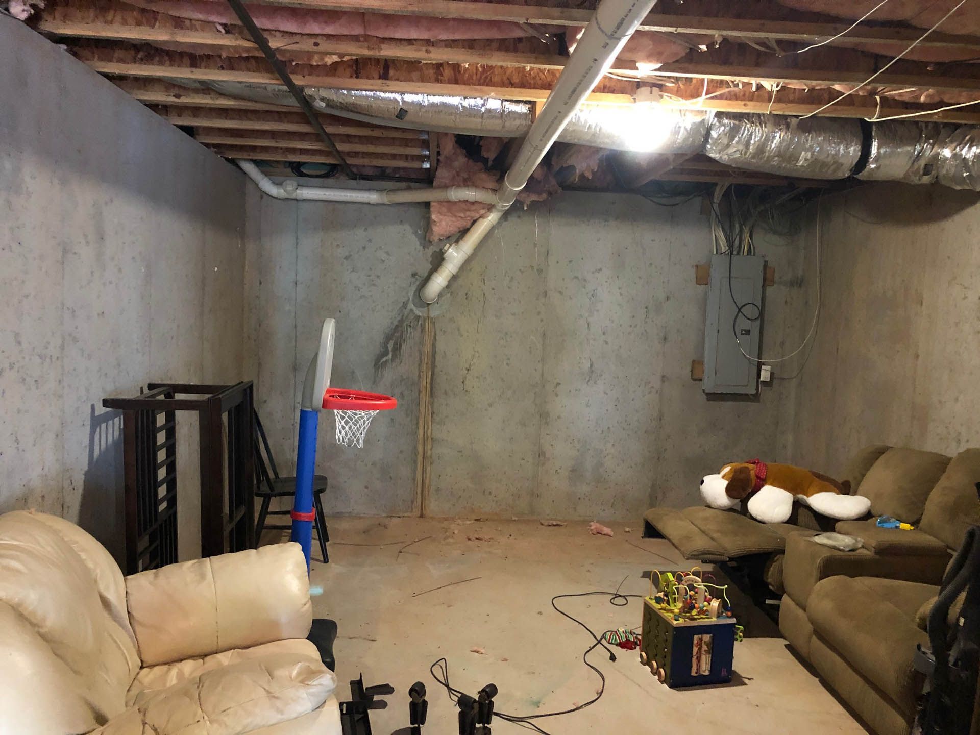A basement play area with concrete walls, exposed ceiling beams, and a basketball hoop. Two couches, toys, and a circuit breaker are visible.
