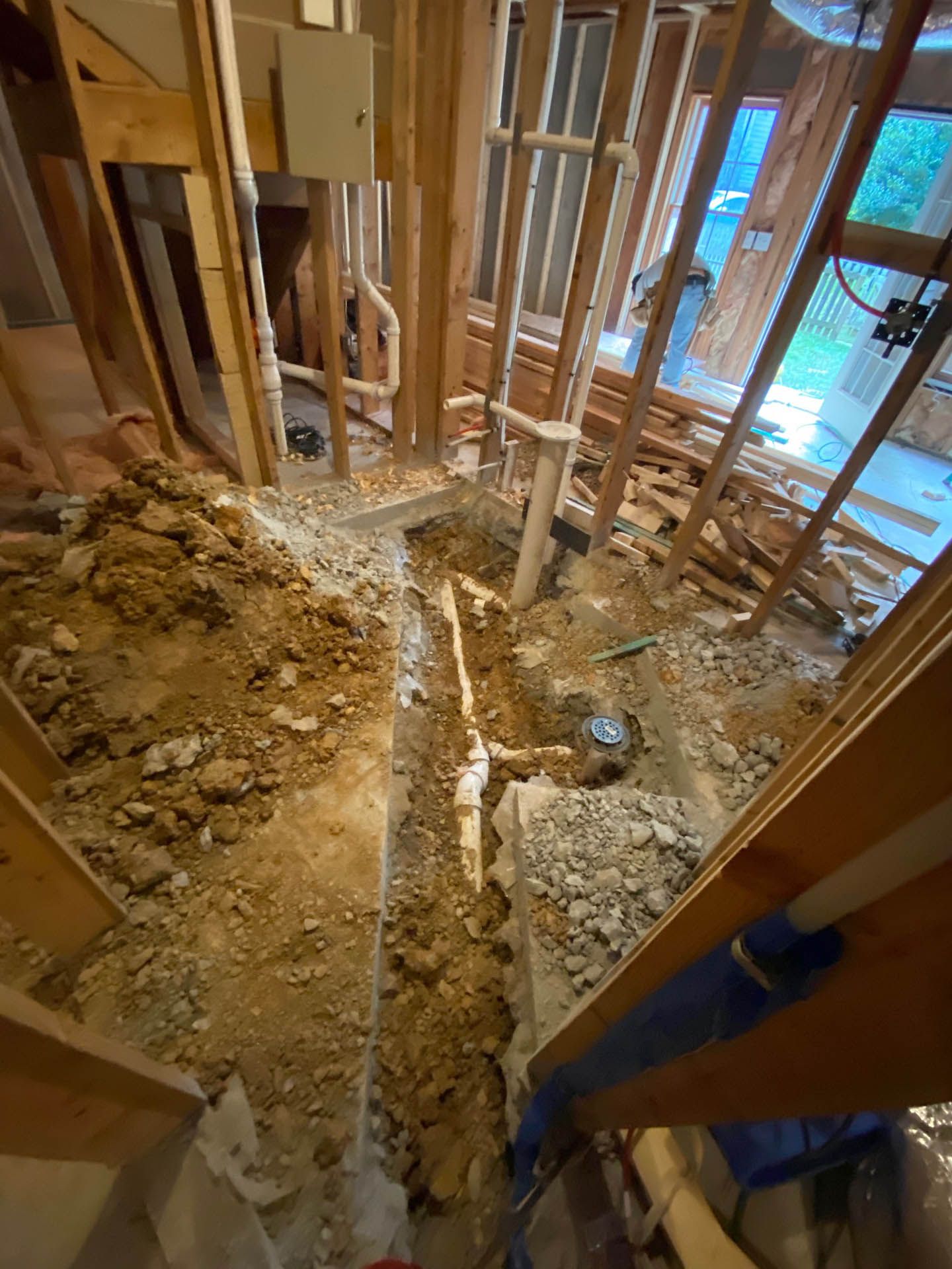 Interior view of a construction site showing exposed pipes, dirt, wooden framing, and unfinished walls.