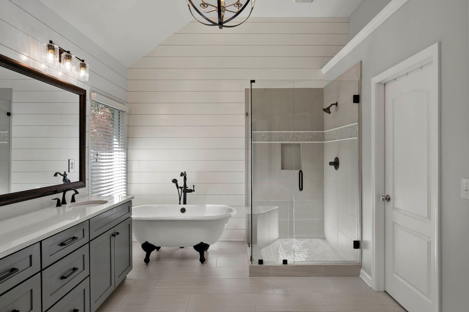 A modern bathroom featuring a gray vanity, white clawfoot tub, and glass shower; light gray tile walls and floor.