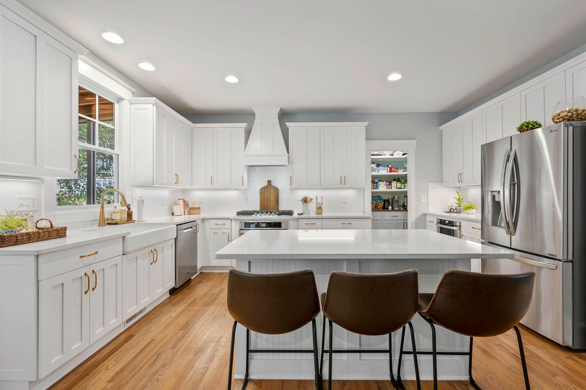 Bright white kitchen with island and three brown barstools, featuring stainless steel appliances and wooden floors.