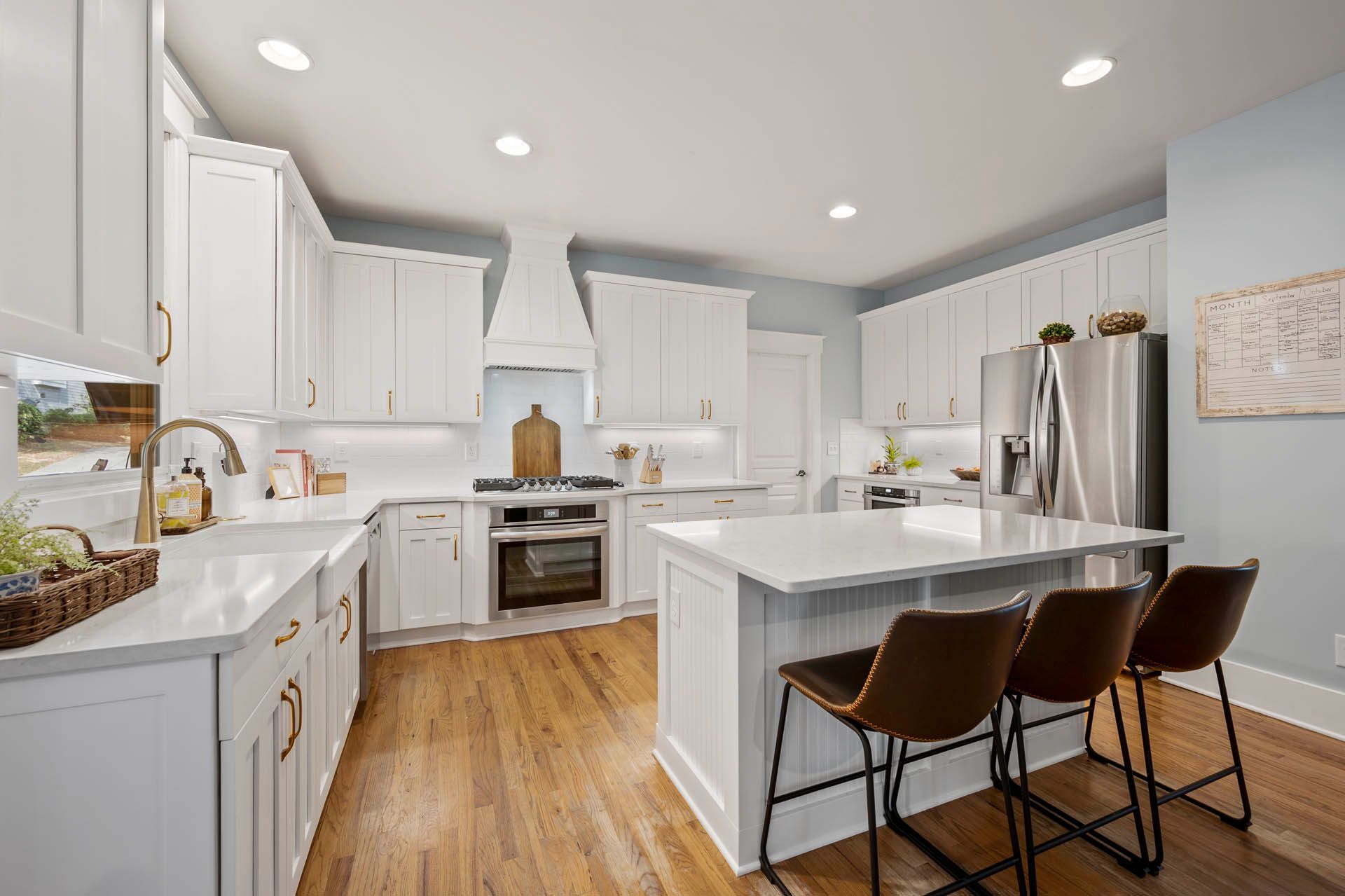 A bright, modern kitchen with white cabinets, marble countertops, and wood floors. Three brown bar stools sit at a small island.