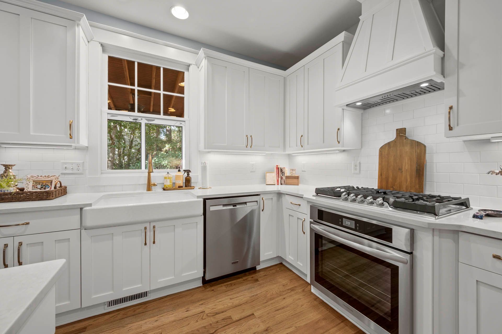 White kitchen with stainless steel appliances, white cabinets, a window, and wooden floors.
