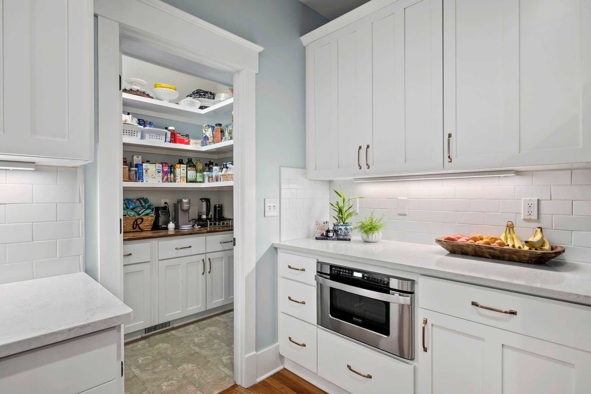 A bright white kitchen with a pantry. Built-in cabinetry, a microwave, and a marble countertop are visible. The pantry has shelves filled with food.