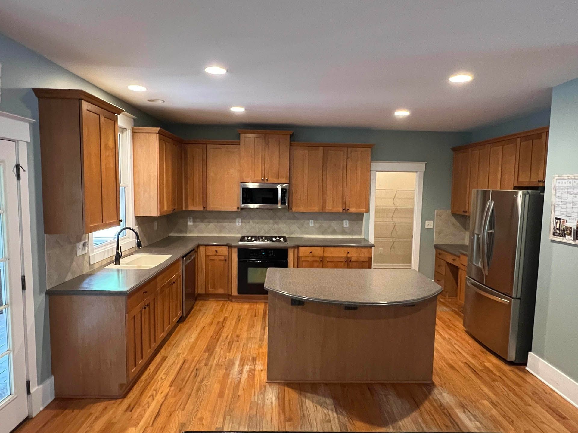 A kitchen with light wood cabinets, gray countertops, and stainless steel appliances. The room has hardwood floors and is painted a pale blue.
