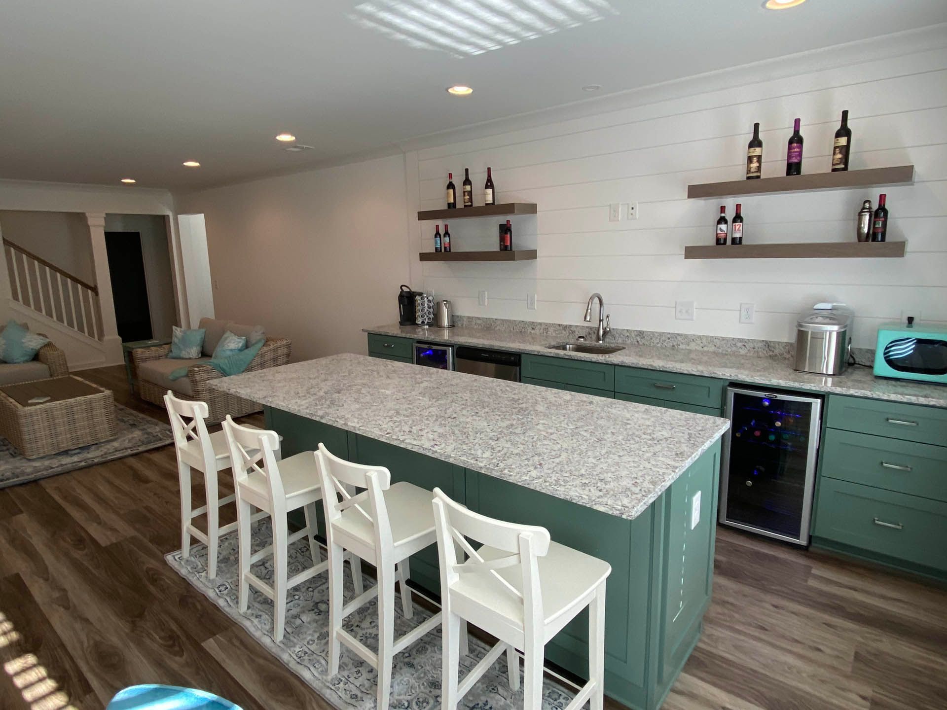 A stylish, teal and white home bar with a granite countertop island and bar stools, shelves with wine bottles, and built-in appliances.