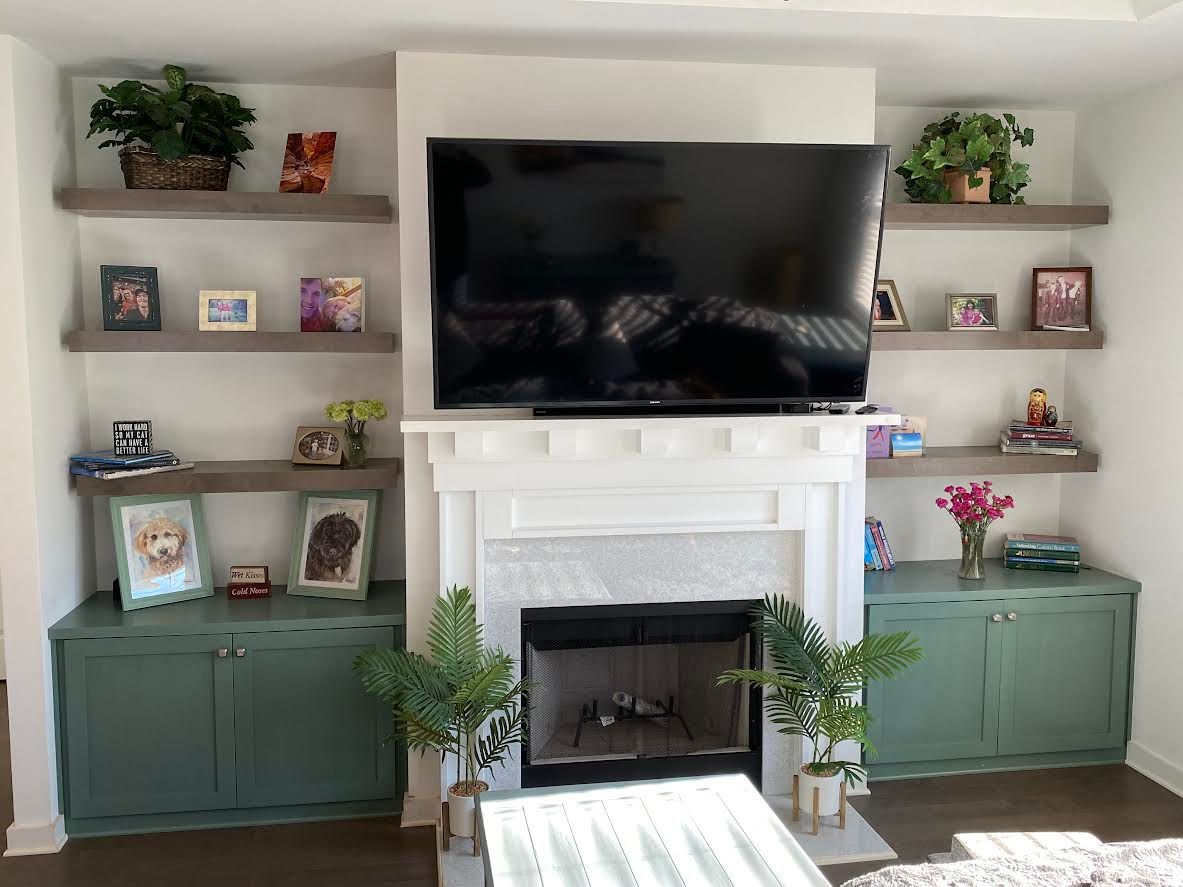 A fireplace flanked by shelves and cabinets, with a TV mounted above it. The cabinets and greenery are a teal color, the shelves are grey, and the walls are white.