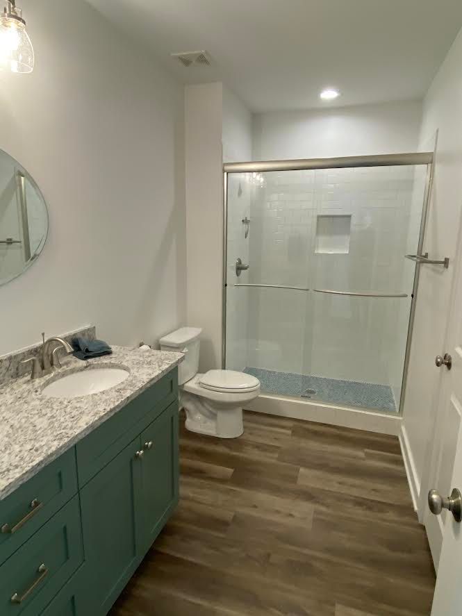 A modern bathroom with teal vanity, wood-look floor, white walls, a glass shower, and a toilet.