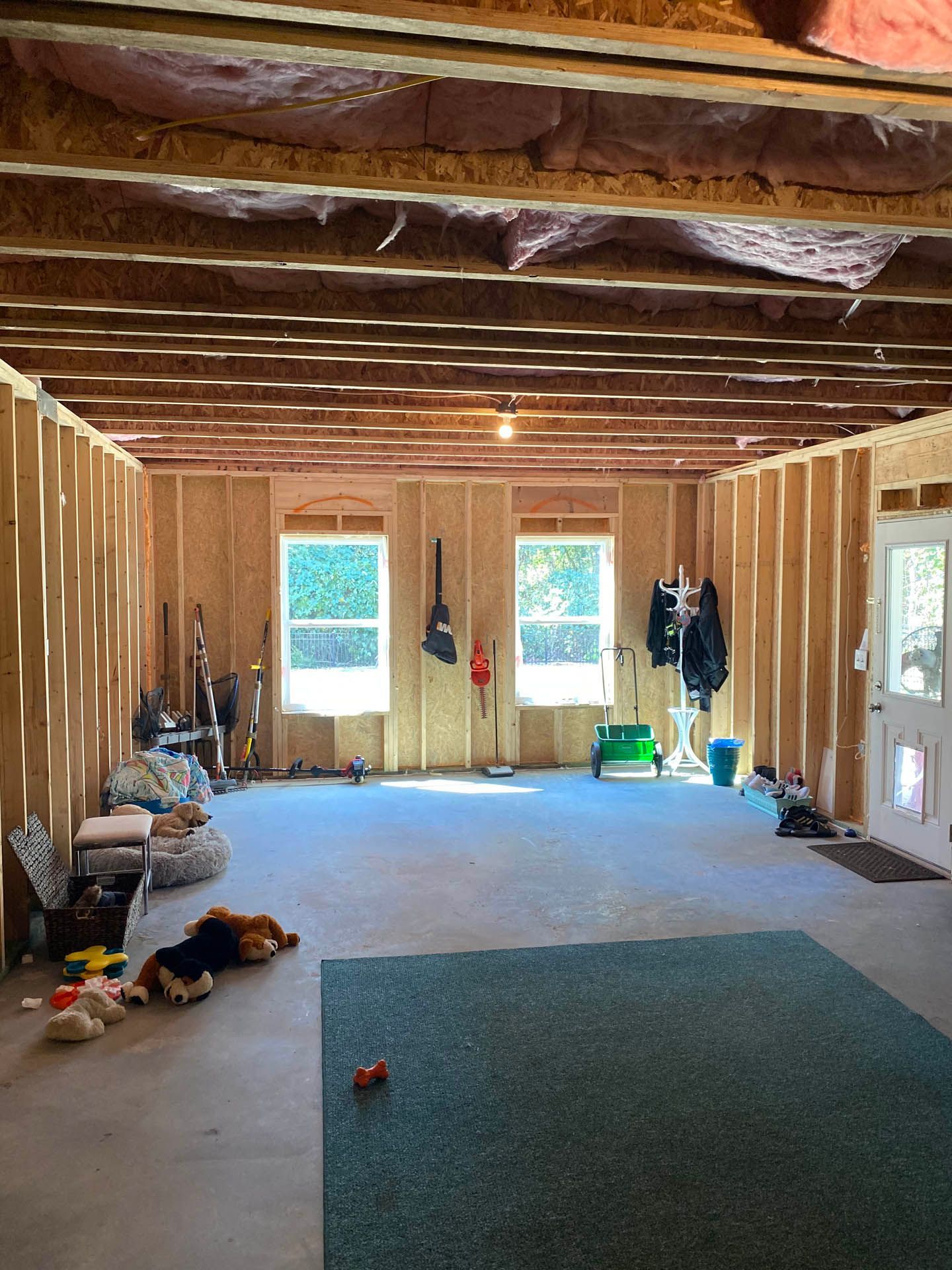 Interior view of an unfinished room with exposed wooden beams, studs, and a concrete floor. Two windows provide light, and a blue rug is on the floor.