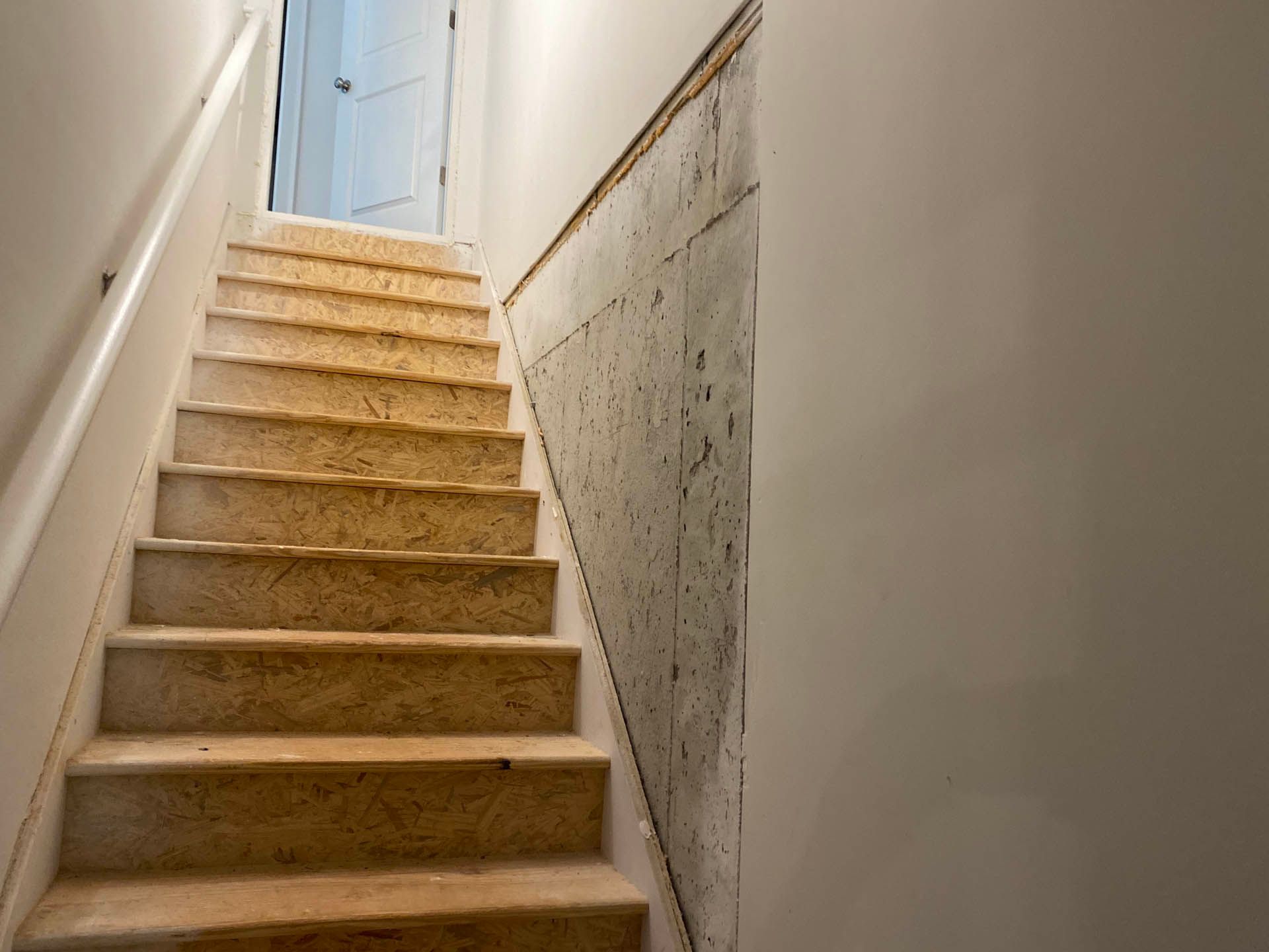 Staircase leading upward, with concrete wall on the right and white painted walls on the left and front. The stairs are worn.