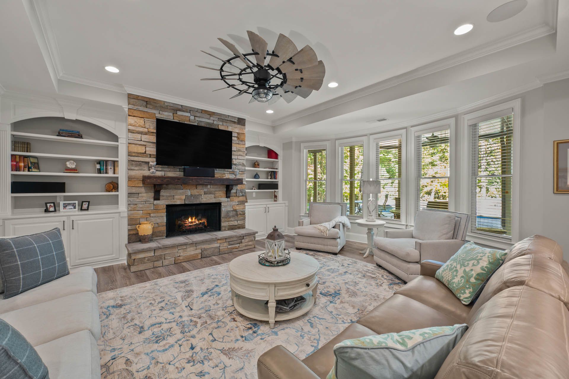 Living room with stone fireplace, built-in shelves, large windows, and beige sectional sofa. A decorative ceiling fan hangs above a round coffee table.