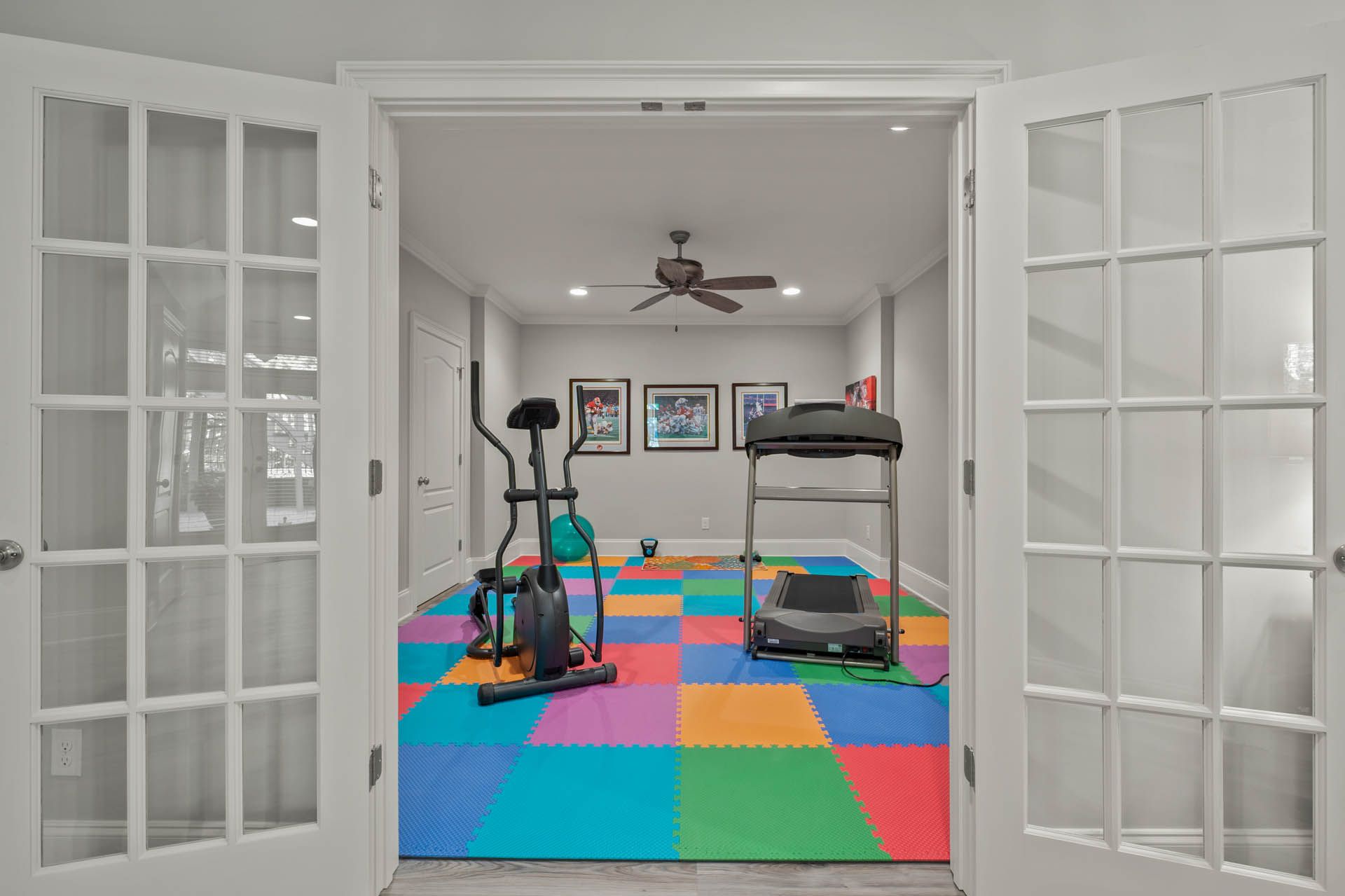 French doors open to a home gym with an elliptical machine, treadmill, and colorful puzzle mat flooring.
