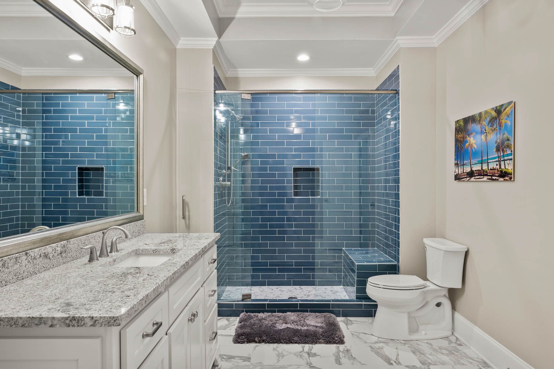 Bathroom with blue tiled shower and wall, white vanity with granite countertop, and white toilet. A painting hangs on the wall.