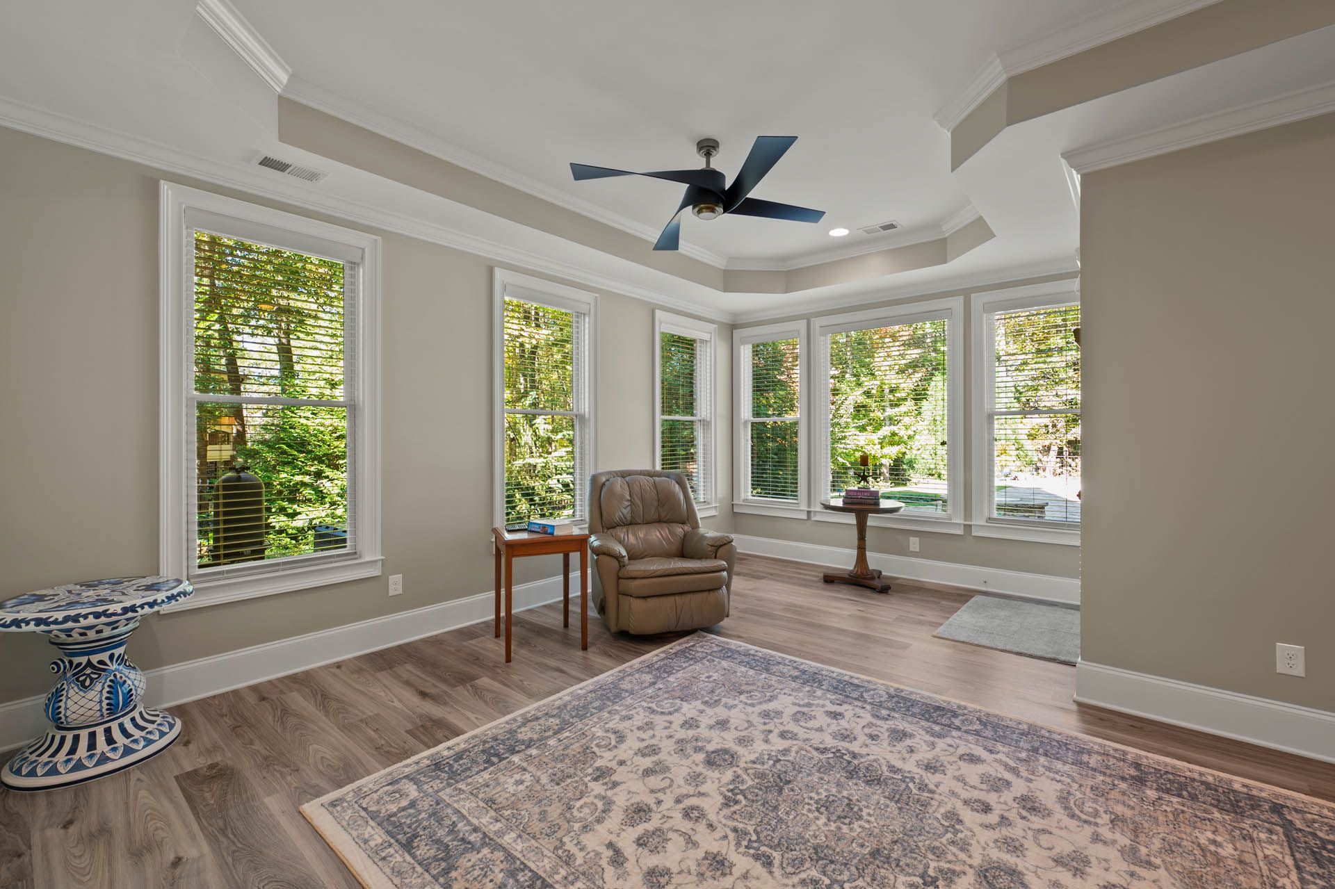A living room with large windows overlooking greenery. Features include a recliner, rug, side tables, and a ceiling fan.