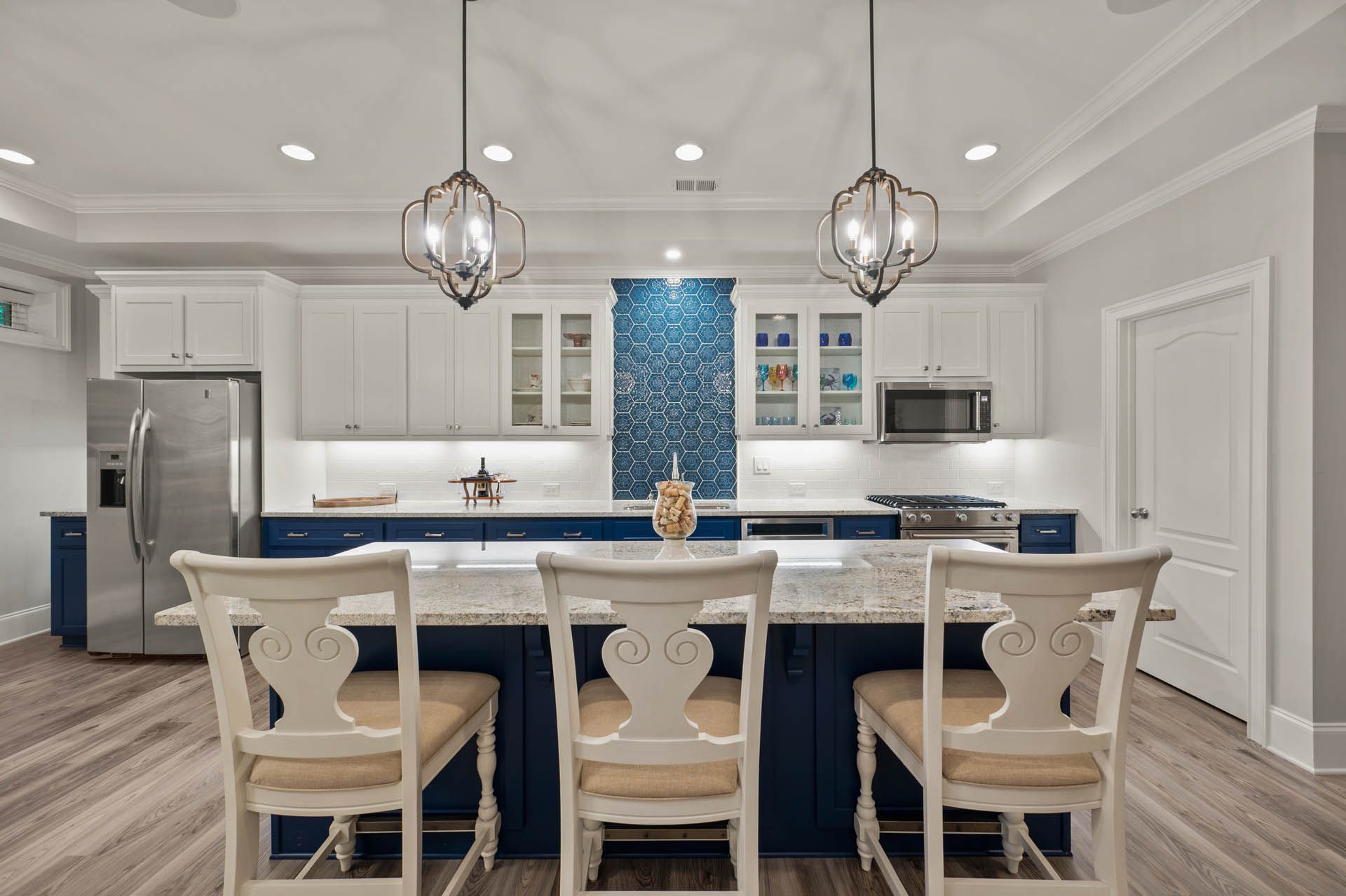 A modern kitchen with blue and white cabinets, a granite island with three white chairs, and ornate light fixtures.