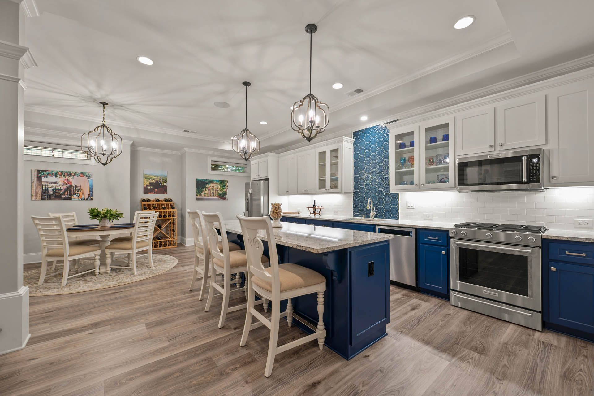 Modern kitchen with white and navy blue cabinets, stainless steel appliances, and a large island. Dining area with round table and chairs is adjacent.