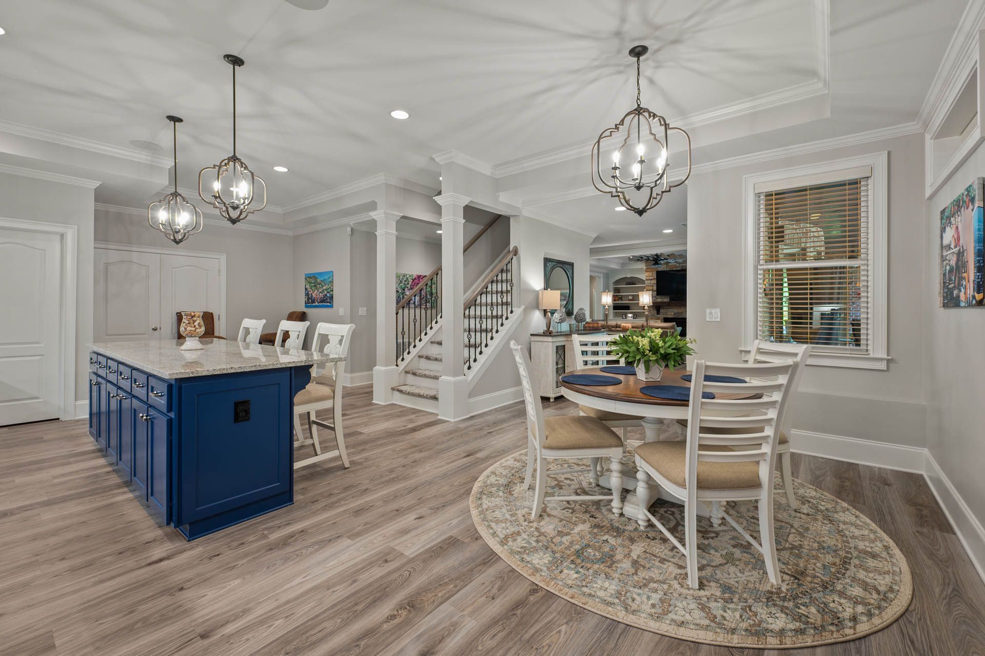 Spacious kitchen with a blue island, dining table, and staircase. Features light wood floors, white walls, and decorative chandeliers.