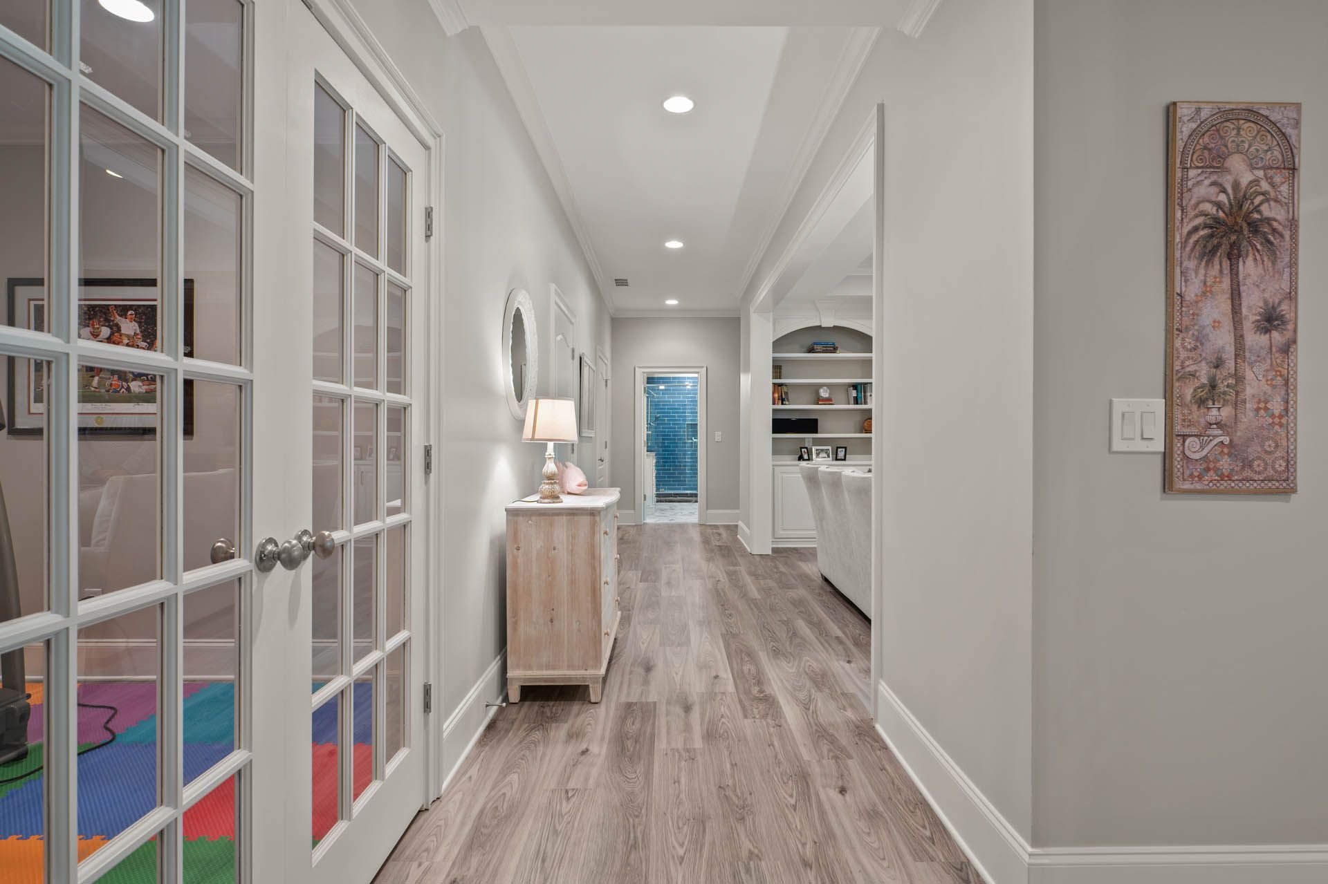 Long hallway with light gray walls and wood-look flooring. French doors on the left lead to a playroom; a console table and art piece adorn the walls.