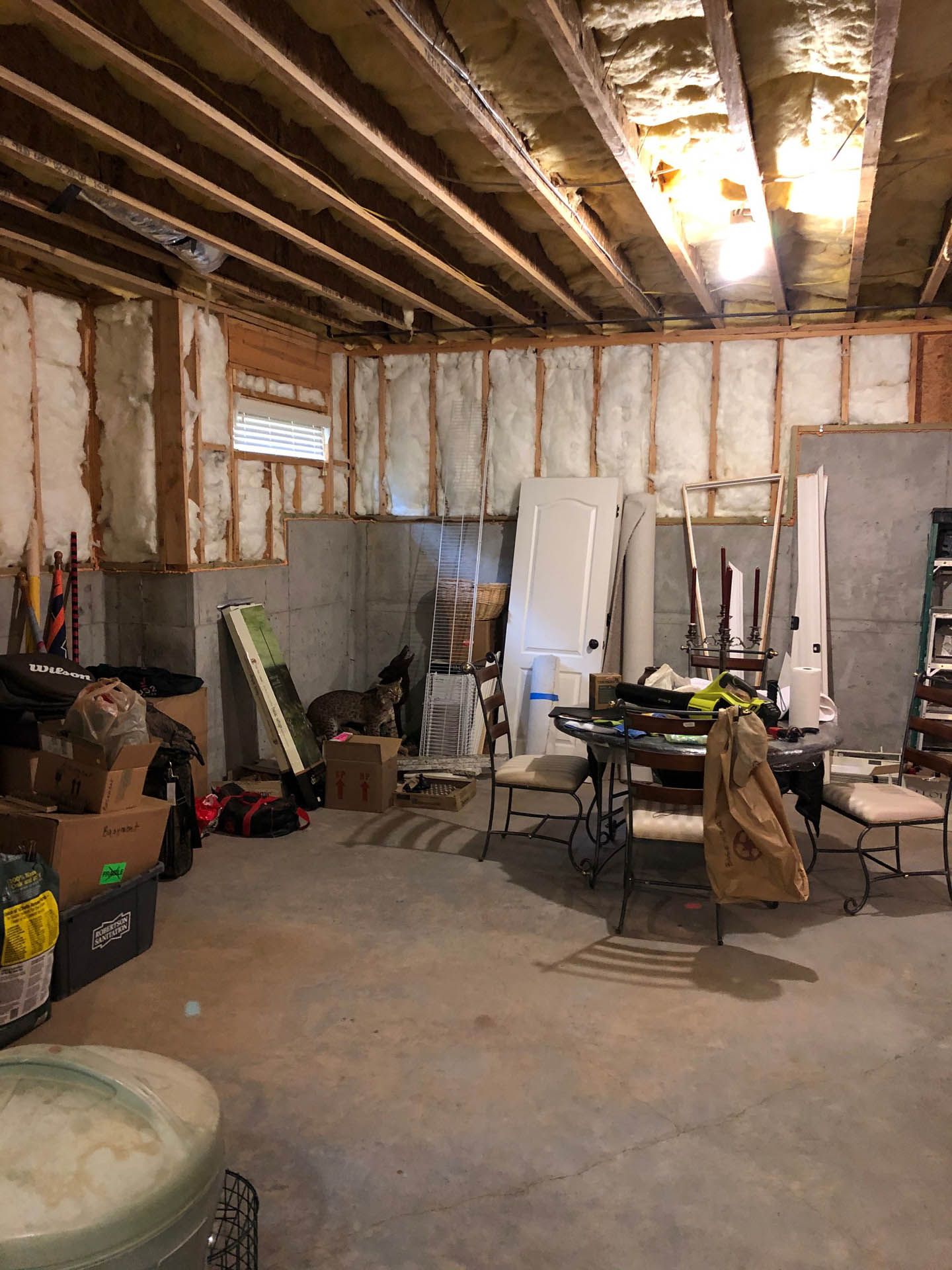 Unfinished basement with exposed insulation and wooden beams. Boxes, furniture, and construction materials are scattered throughout.