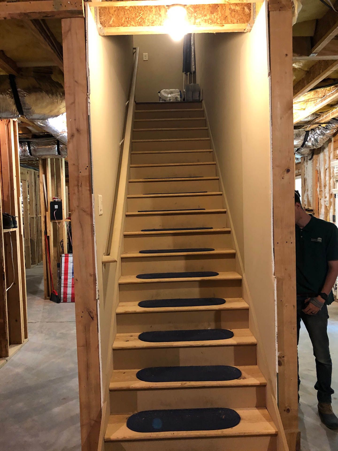 A wooden staircase leads upwards, with a person partially visible on the right.  The steps have black treads. The walls are beige and light-colored wood.