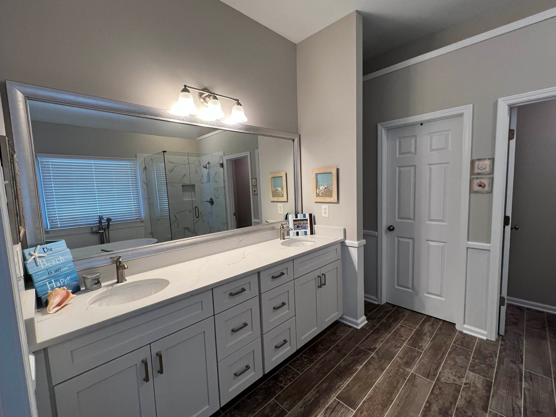 Bathroom with gray walls, white vanity with a large mirror, and dark wood-look tile flooring. A closed door and a partially visible shower are also present.