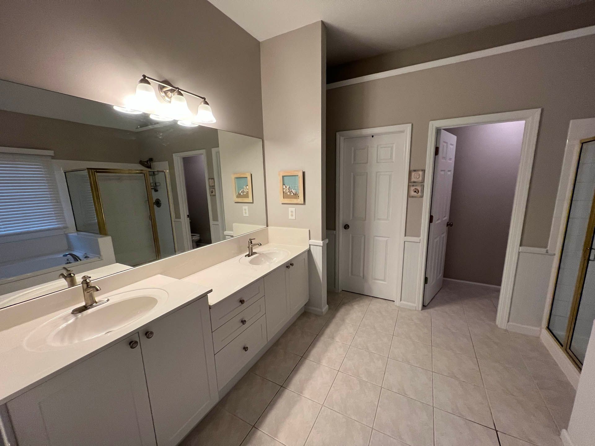 Bathroom with white cabinets, countertops, and tiled floor. A large mirror above the double sink reflects a bathtub and window. Two doorways are also visible.