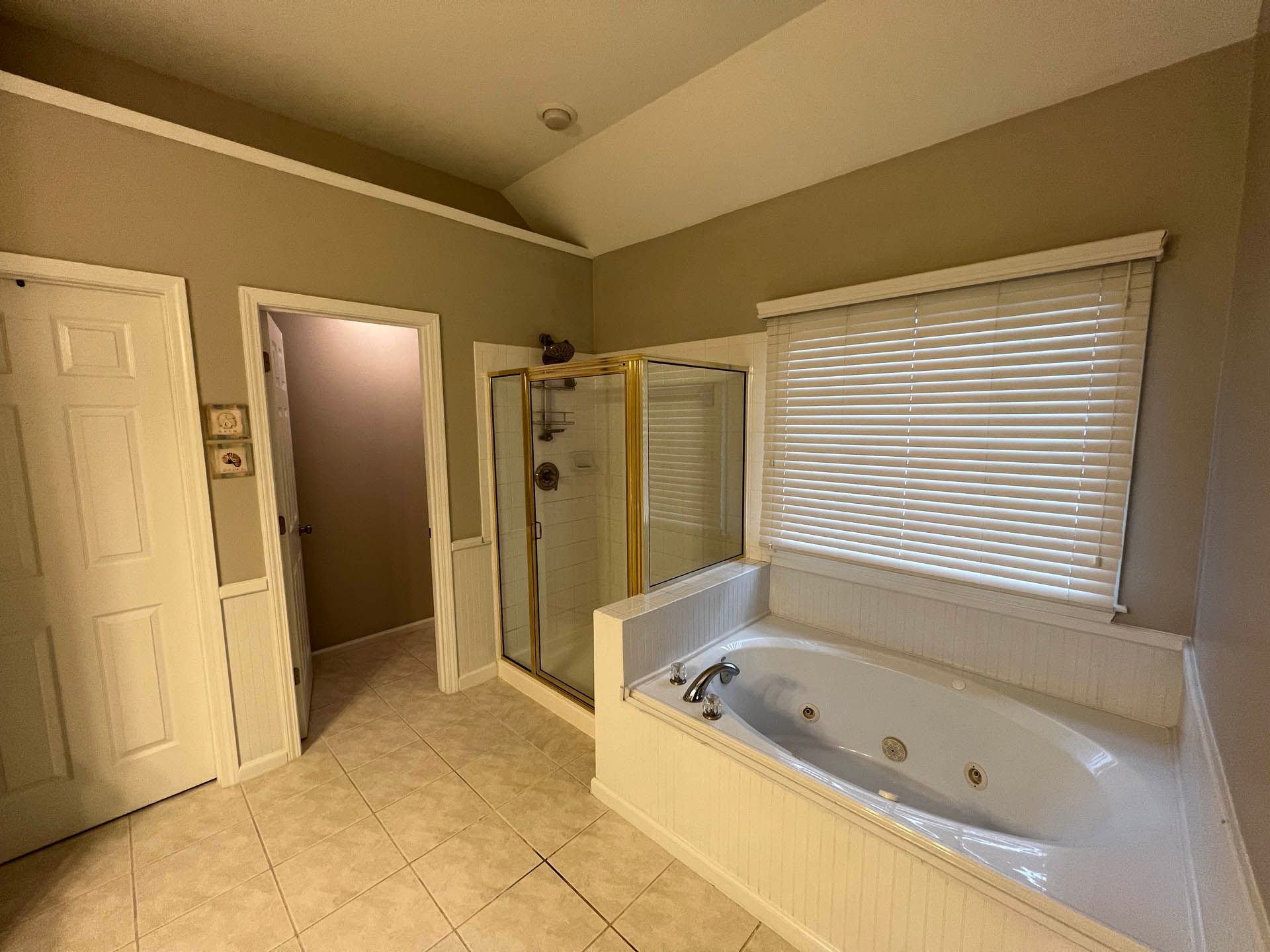 Bathroom with a jacuzzi tub, shower, and doorway. Beige walls and tile flooring. A window is covered with blinds.