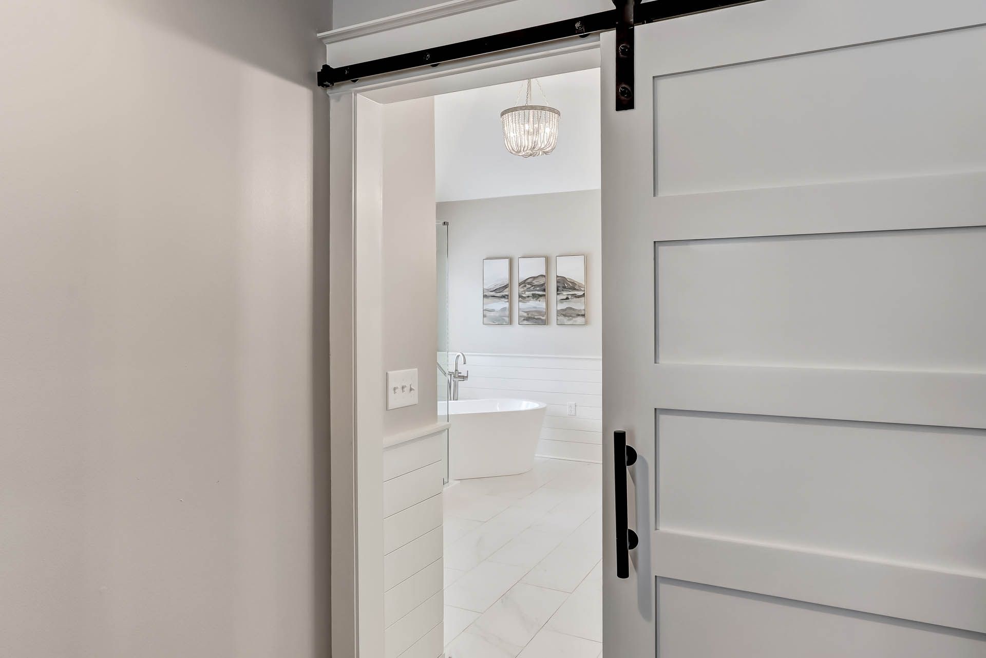 White barn door open to a light-filled bathroom with a chandelier and artwork.