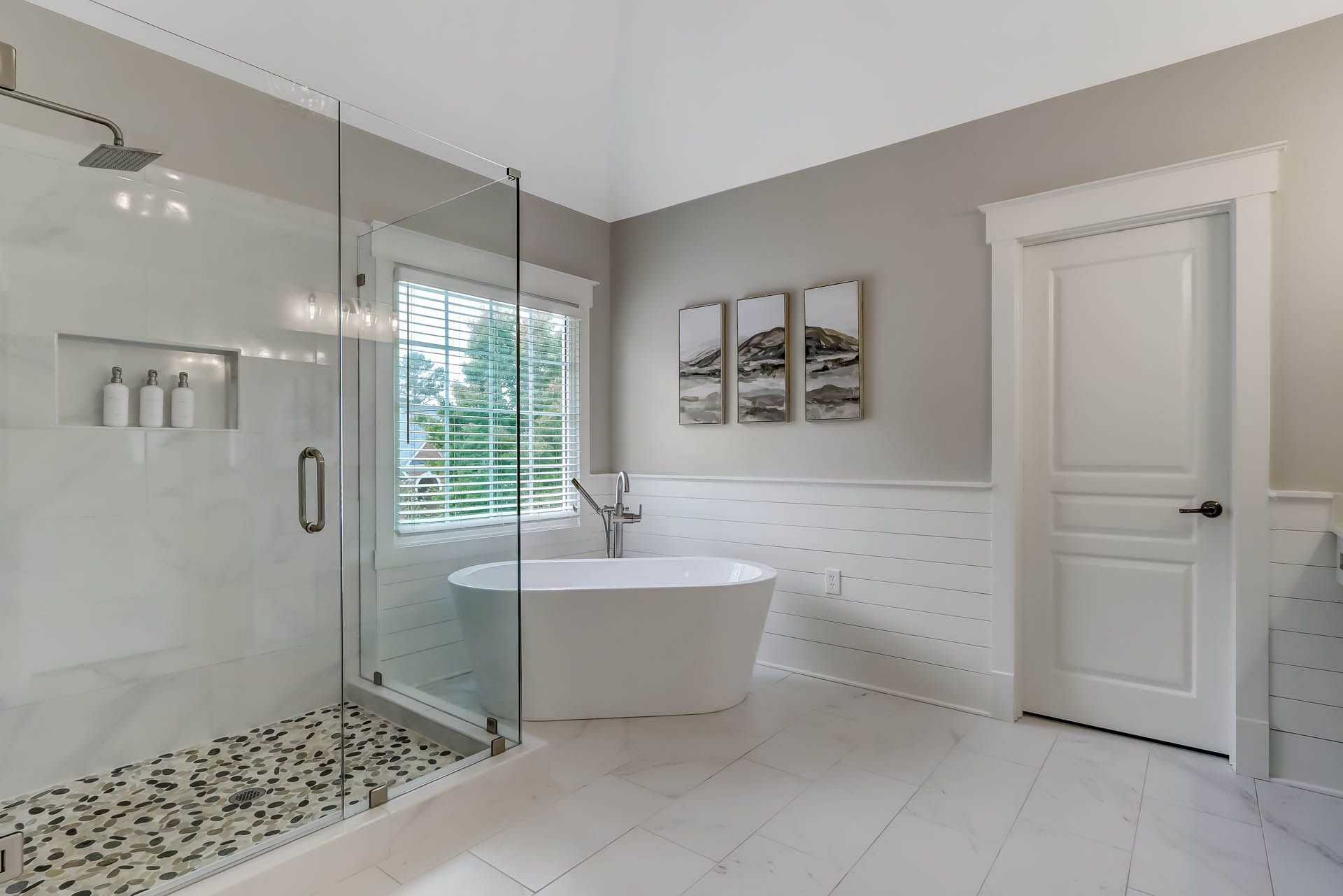 Spacious modern bathroom with a glass-walled shower, freestanding tub, white door, and artwork over the tub.