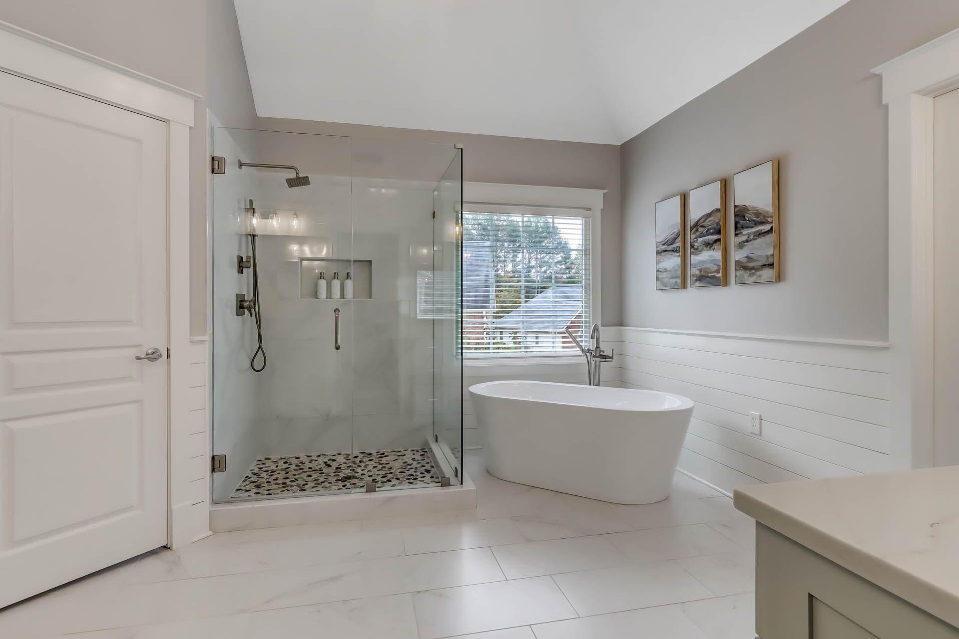 A bright bathroom with a glass shower, freestanding tub, and white tile. Soft gray walls and white trim.