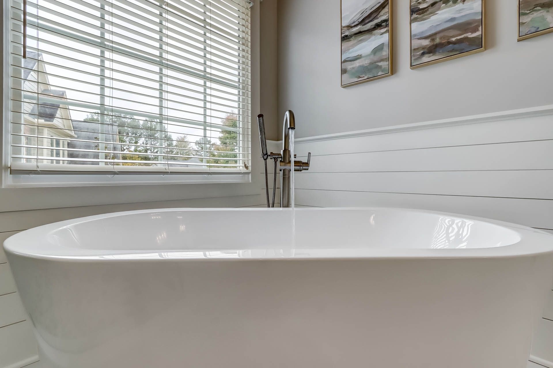 A white soaking tub sits in a bright bathroom with a window and artwork. The walls are white and the fixtures are chrome.