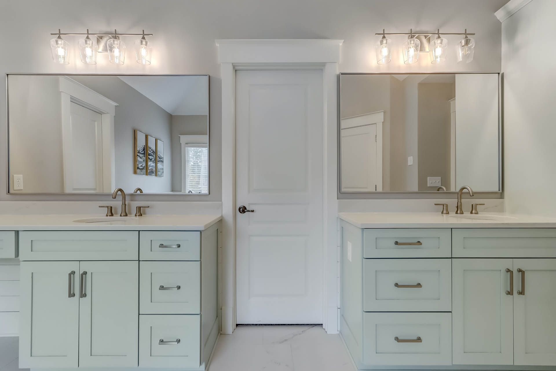 Two bathroom vanities with mirrors, pale blue cabinets, and bright lighting flanking a white door in a gray-walled bathroom.