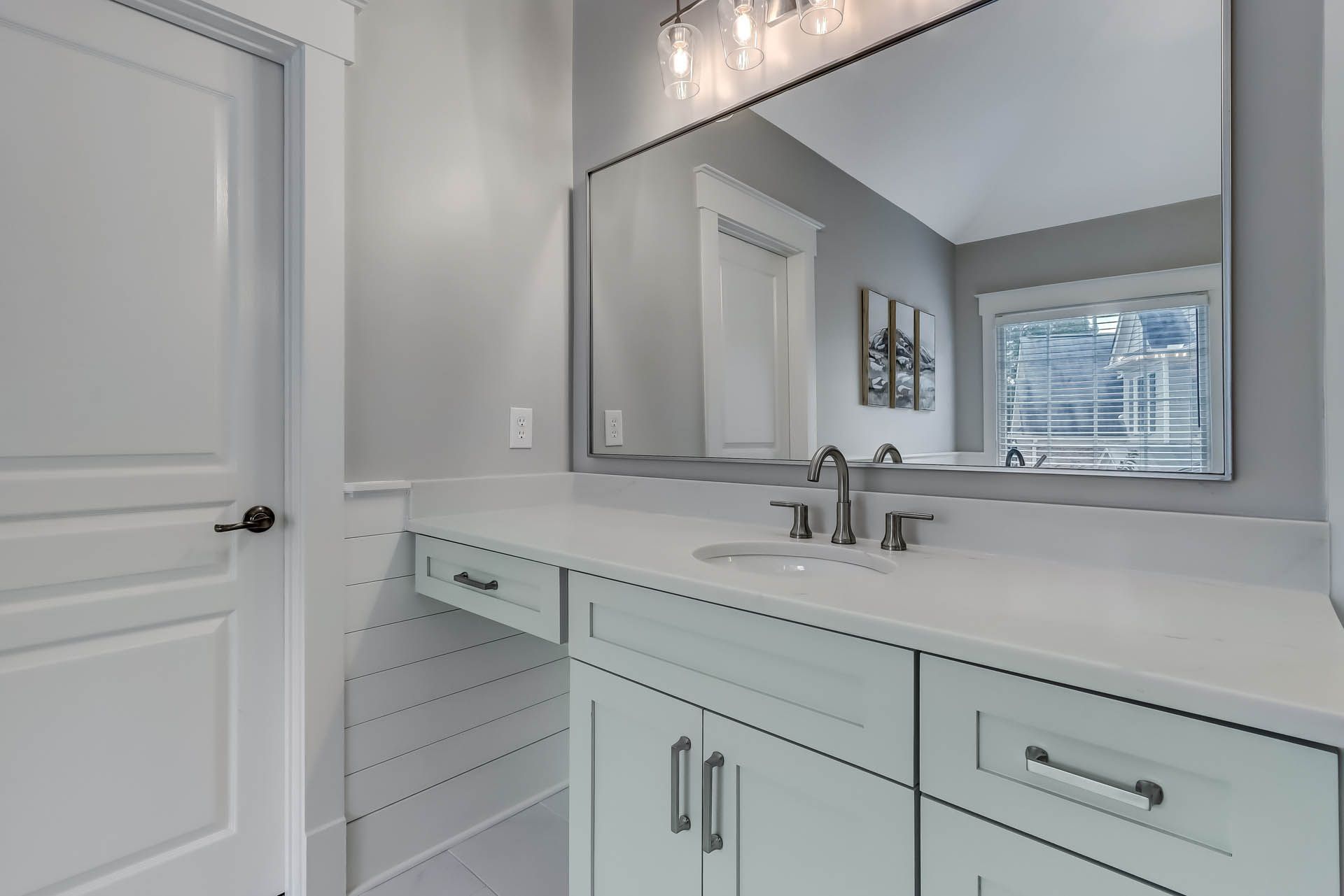 Bathroom with white cabinets, countertop, and door. Large mirror reflects window and light fixture.