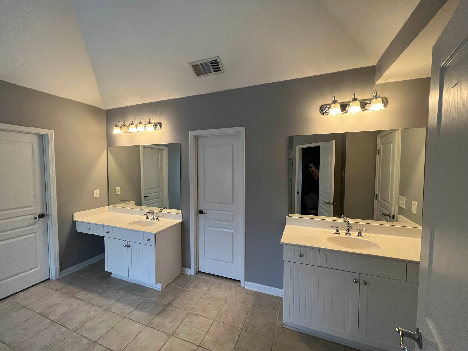 A spacious gray-walled bathroom with two white vanities, mirrors, and overhead lighting. Features include a tile floor, white doors, and a vaulted ceiling.