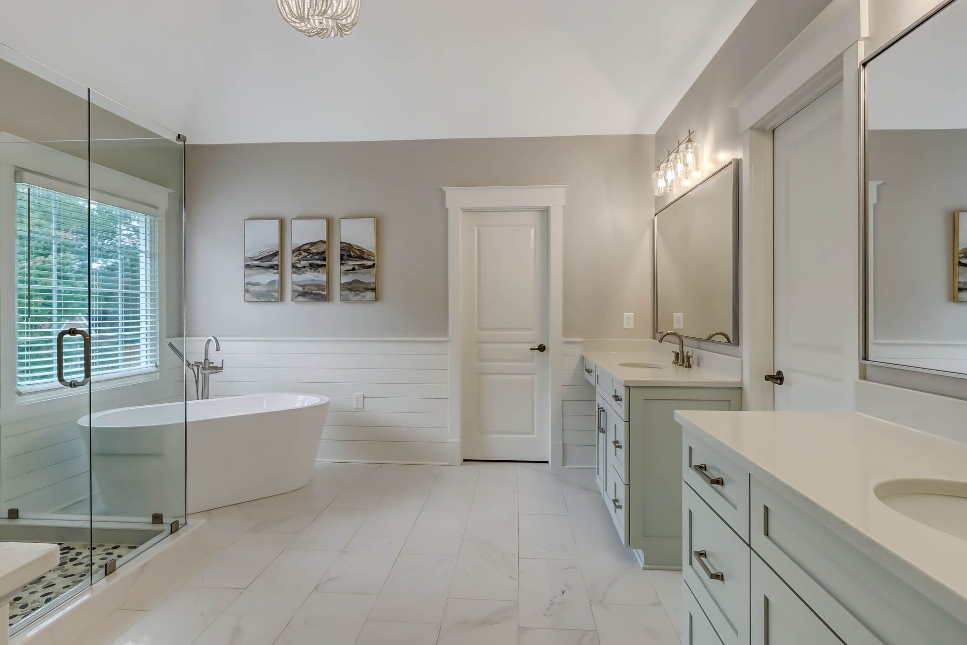Spacious, modern bathroom with a white freestanding tub, glass shower, and double vanity. Gray walls, white trim, and marble-look flooring.