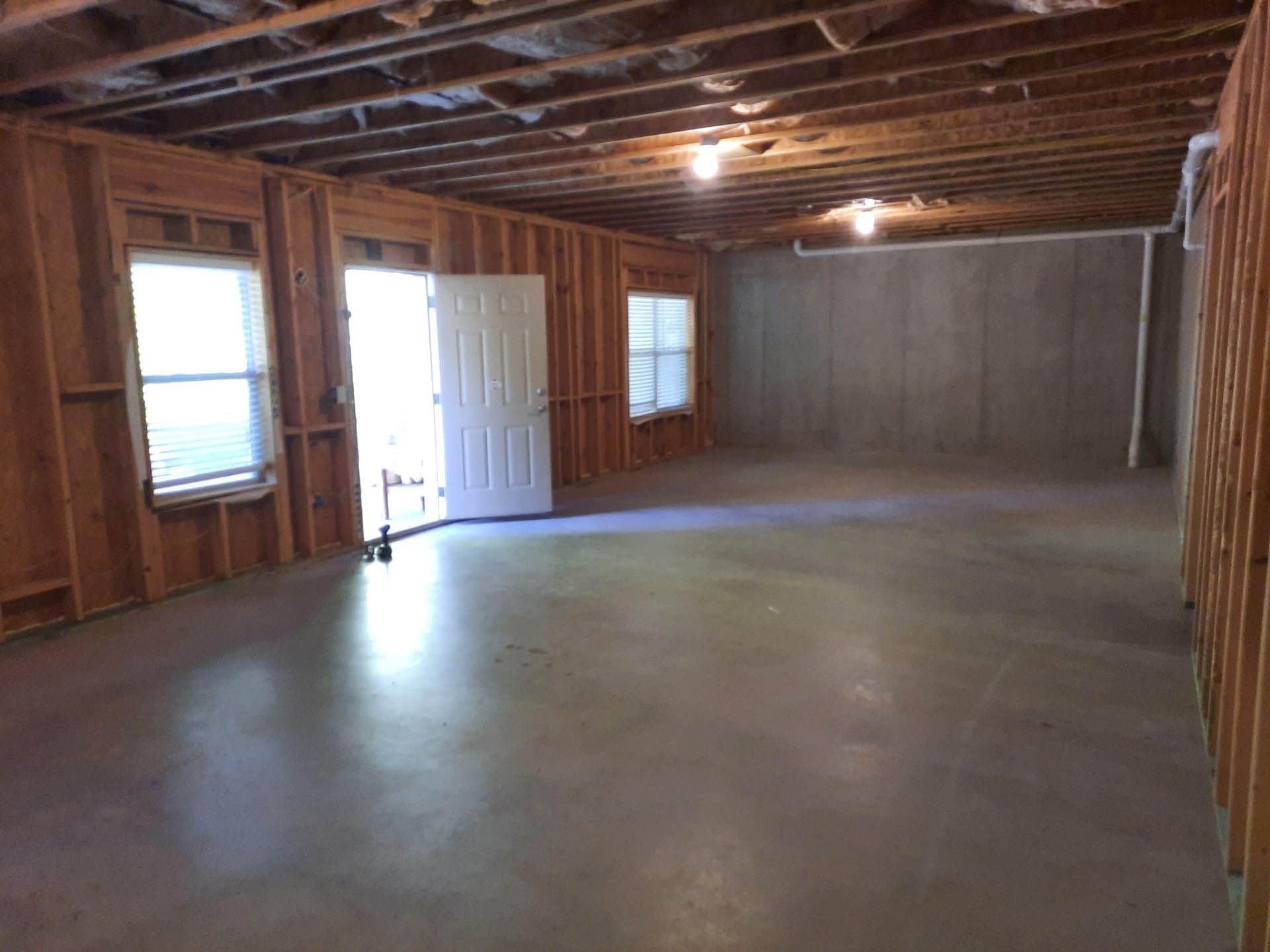 An unfinished basement with a concrete floor, wooden framing, and a door and windows. The room is empty.