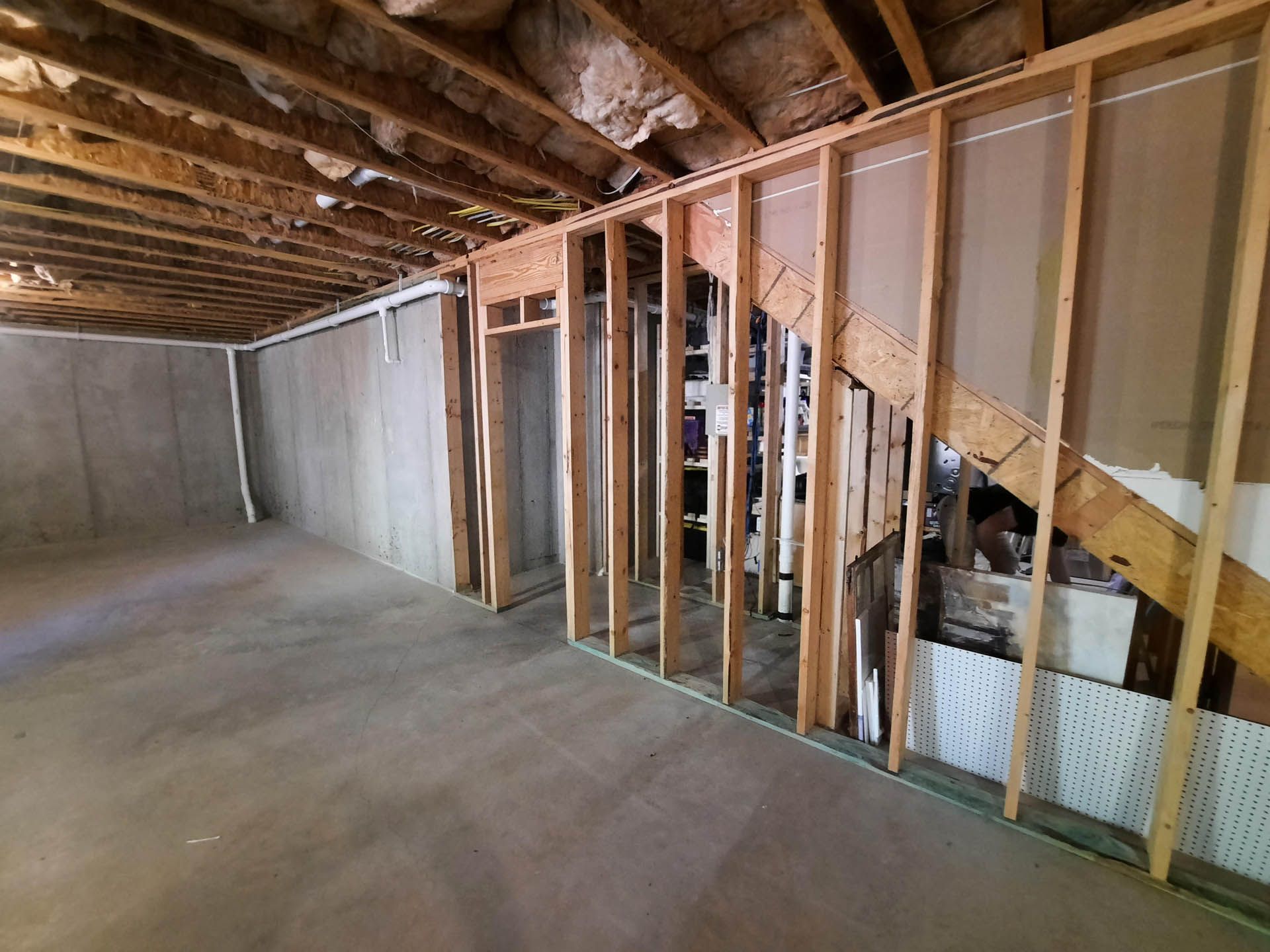 Basement interior under construction. Framing for walls and stairs visible against concrete foundation, with exposed ceiling beams.