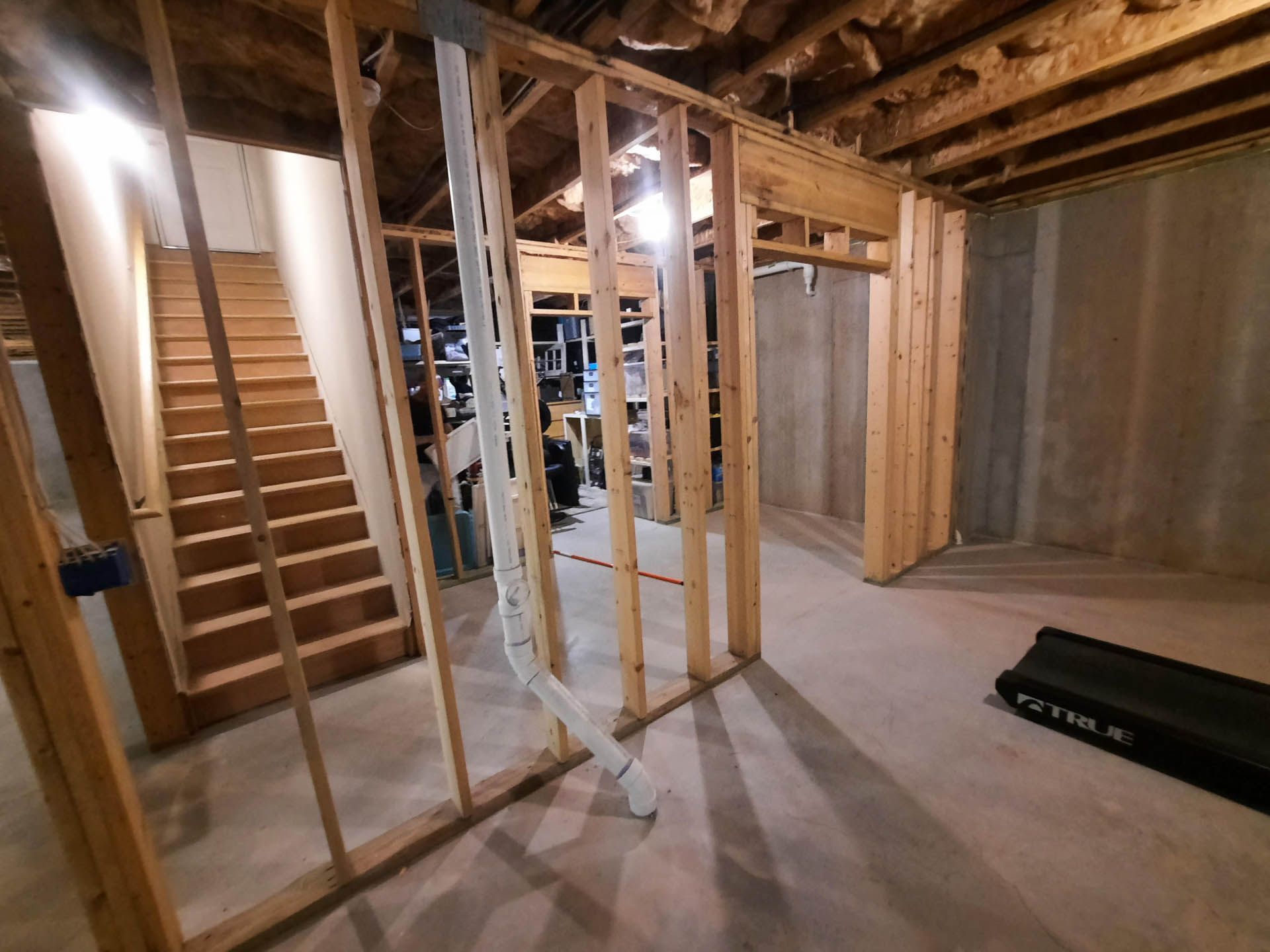 Basement under construction with exposed wooden framing, stairs, and concrete walls. Ductwork hangs from the ceiling.