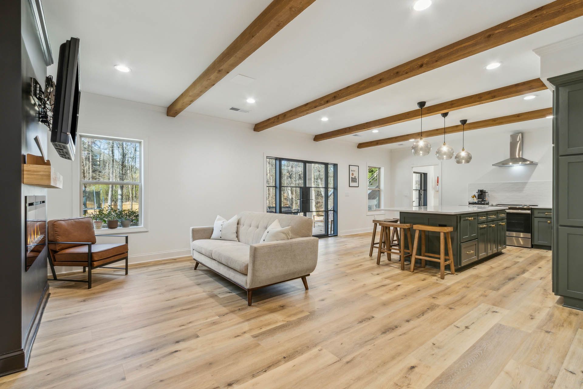 Open-concept living space with a modern kitchen, light wood floors, and exposed wooden beams. A living area features a neutral sofa and a window overlooking a yard.