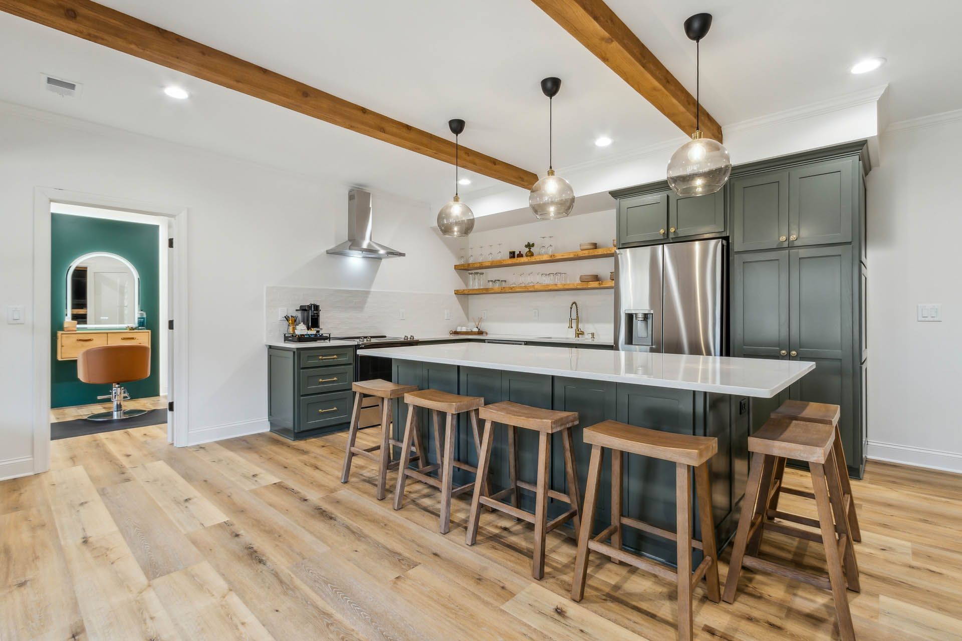 Modern kitchen with a large island and stools, dark green cabinets, and wooden beams. A doorway with a vanity is visible.