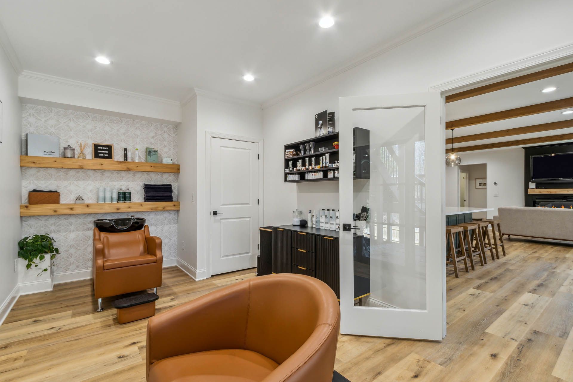 A hair salon interior with wooden floors, tan leather chairs, and white walls. Products sit on floating shelves.