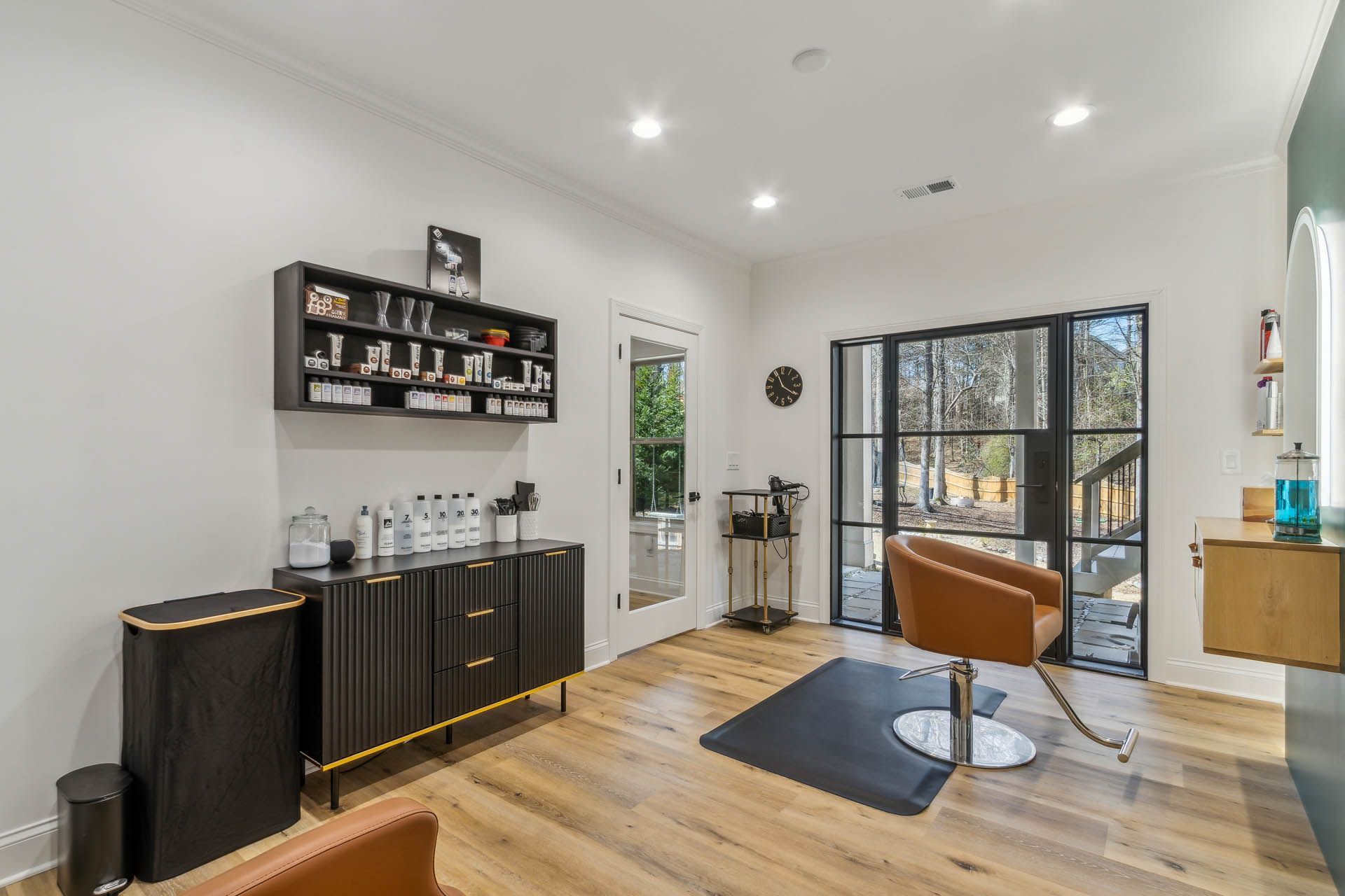 A modern hair salon interior with a styling chair, cabinet, and large windows overlooking an outdoor space.