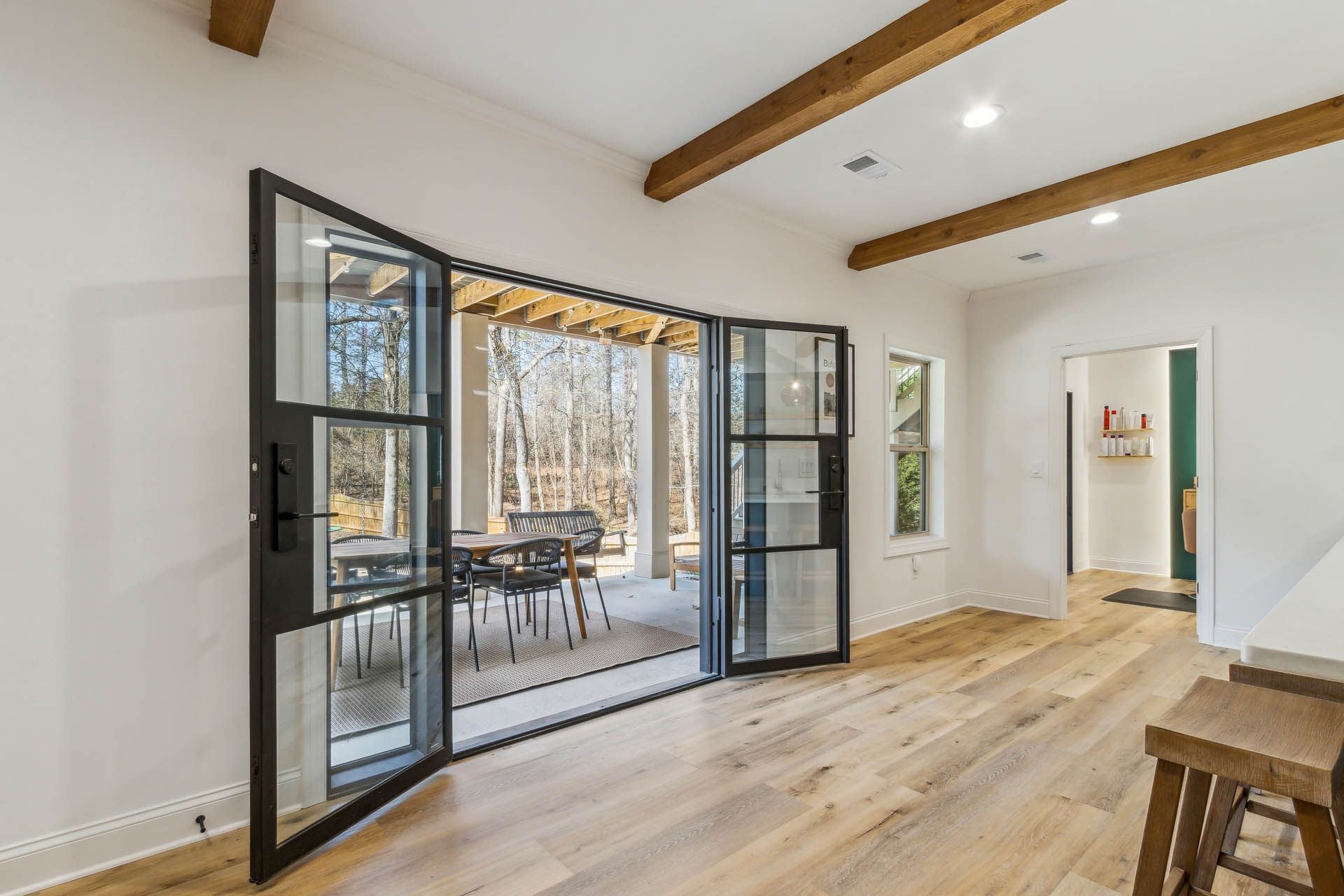 Open, black-framed doors reveal a patio with a dining table. Wooden beams and floors contrast against white walls.