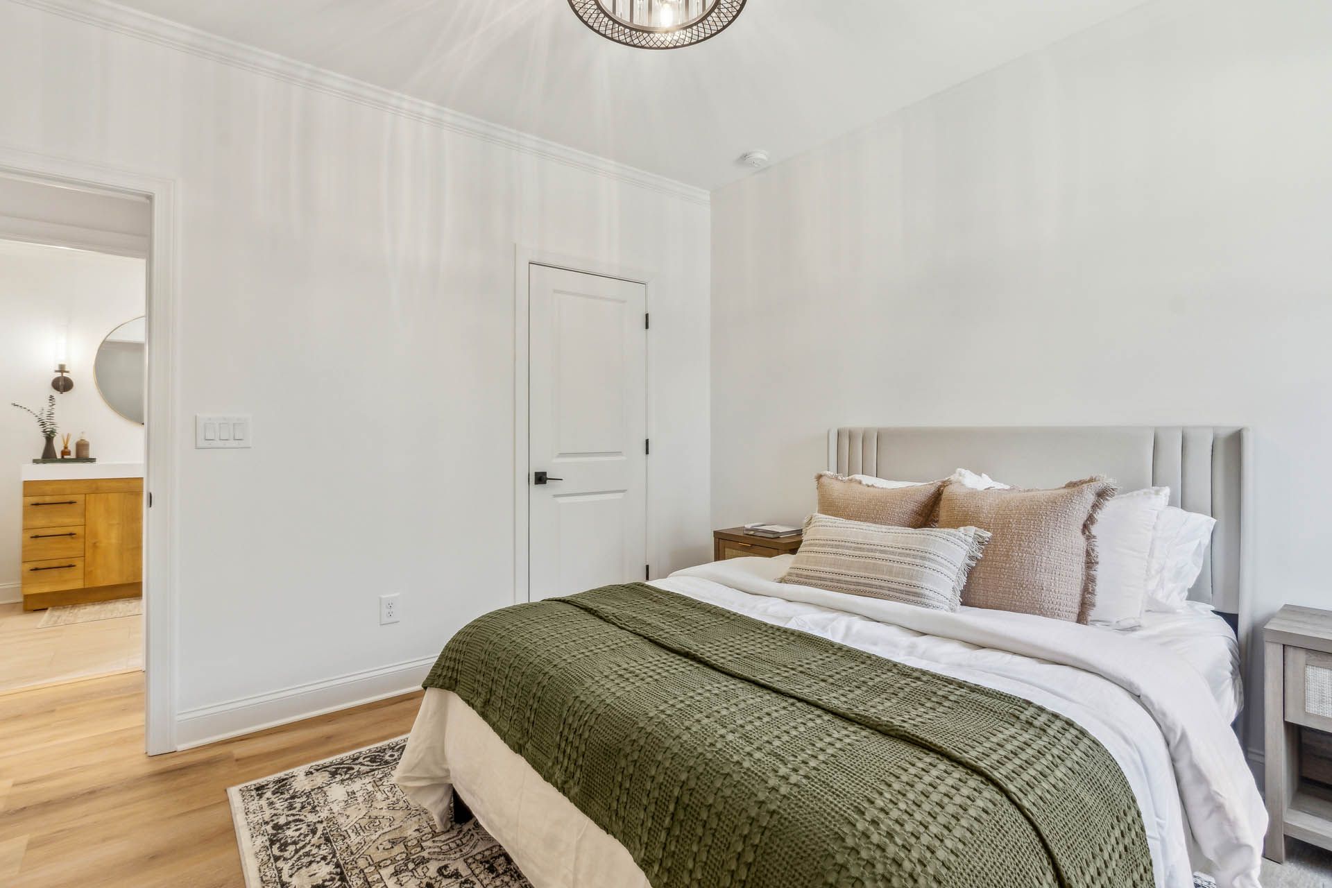 A bedroom with a bed covered in a green blanket, beige pillows, and a wooden nightstand. A door leads to a bathroom.